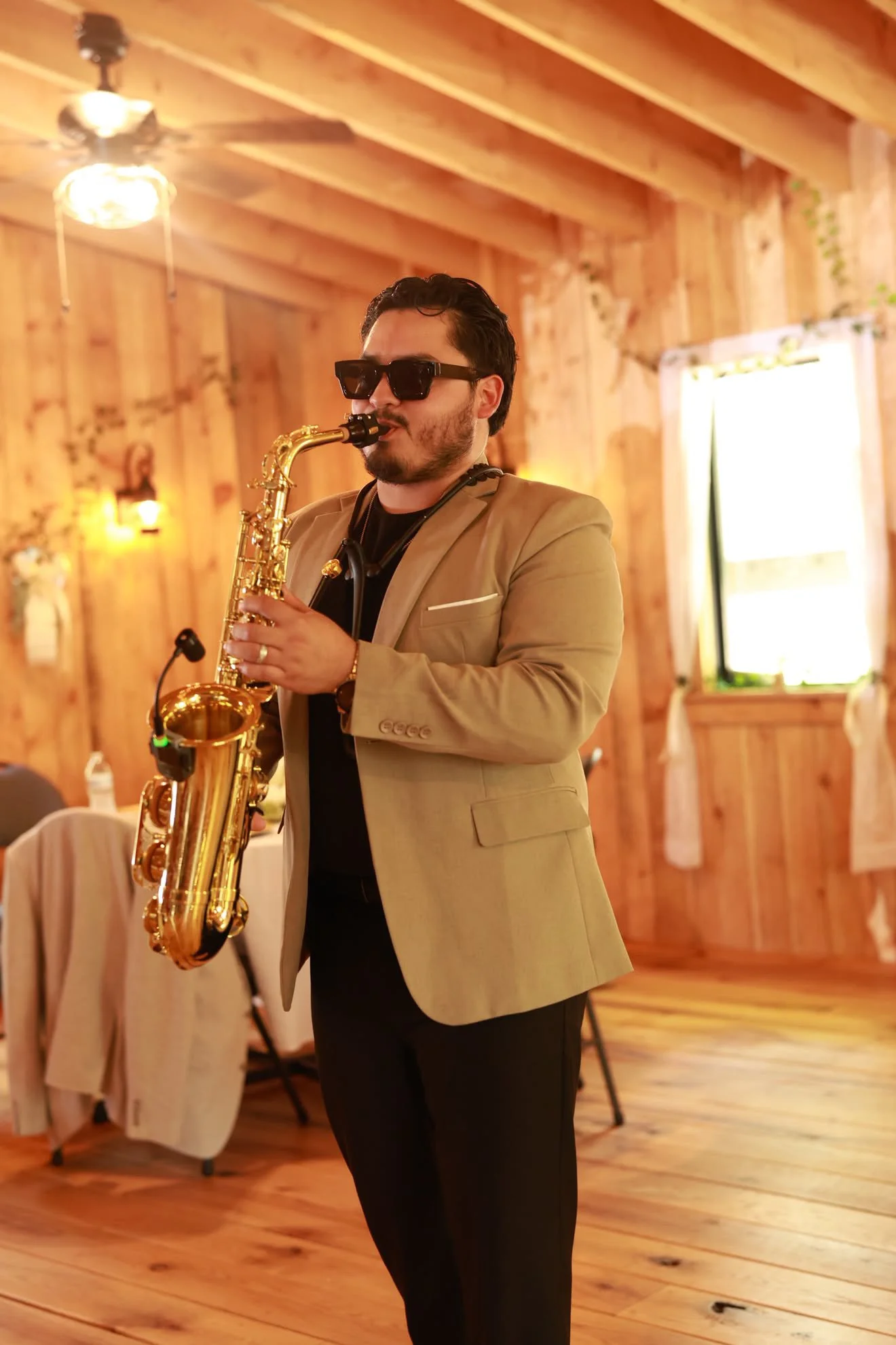 A man with dark hair, beard, wearing black sunglasses, a beige blazer, and black pants, playing a gold saxophone in a wooden room with a ceiling fan and a window with white curtains.