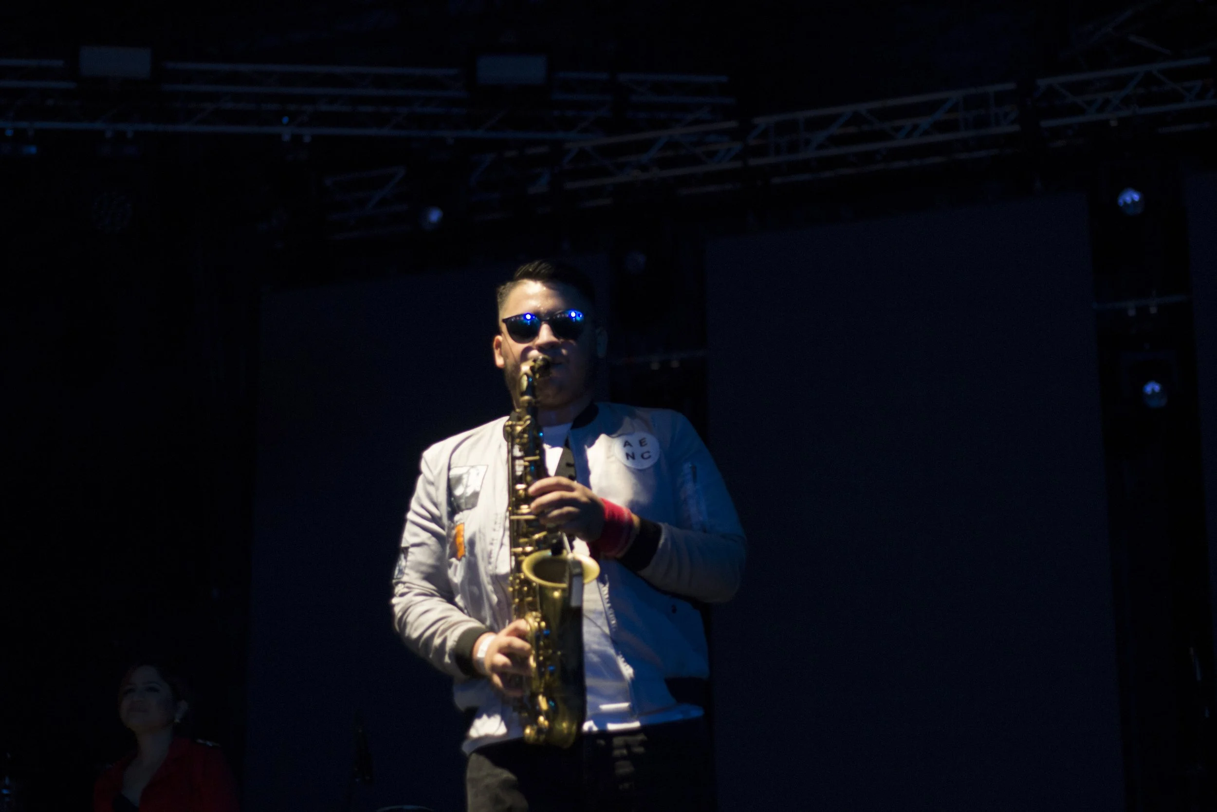 A saxophone player on stage wearing sunglasses and a white jacket, illuminated by stage lights with a dark background.