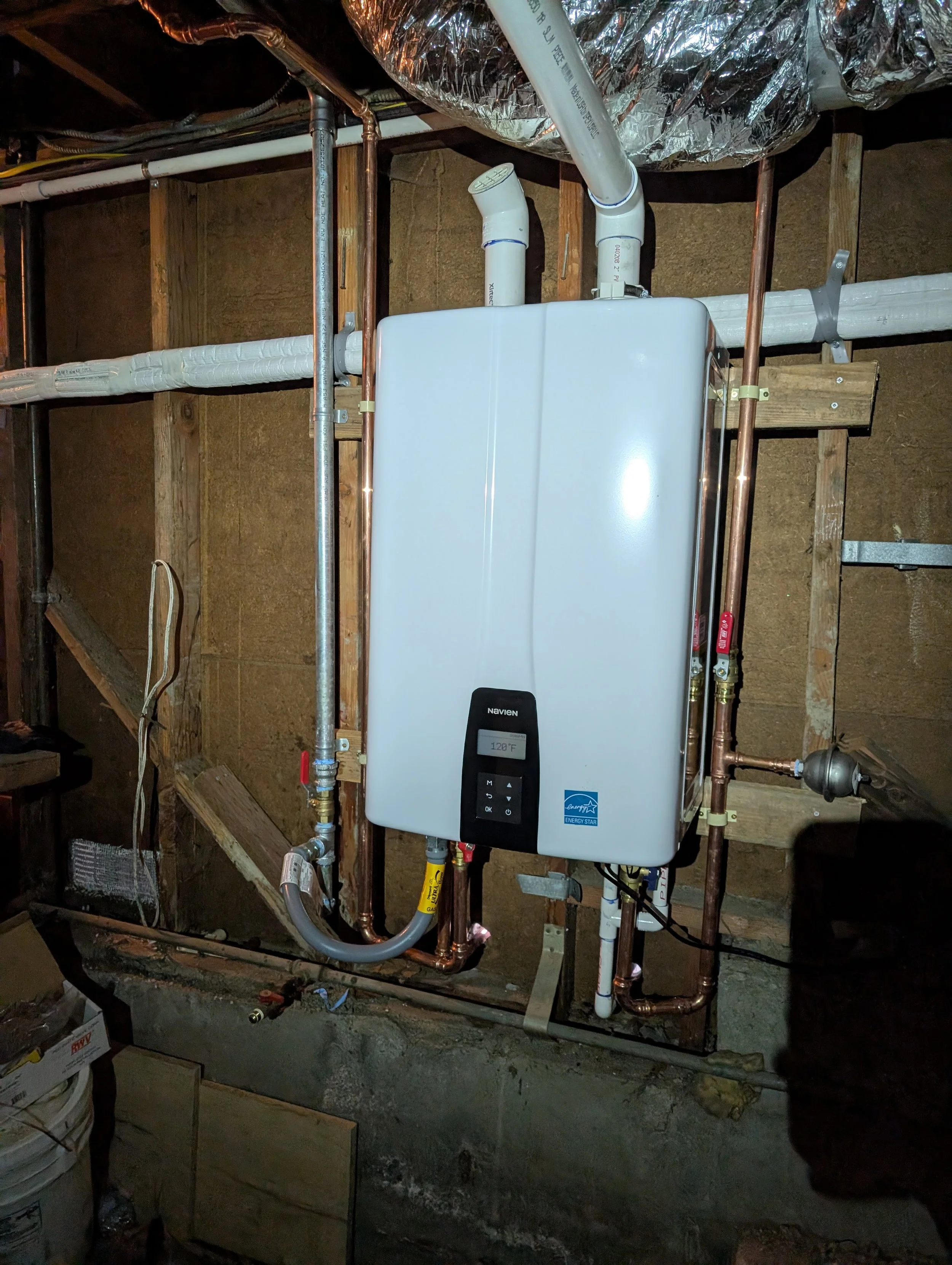 A tankless natural gas water heater installed in a basement with exposed copper and plastic pipes, surrounded by unfinished wooden framing and insulation.