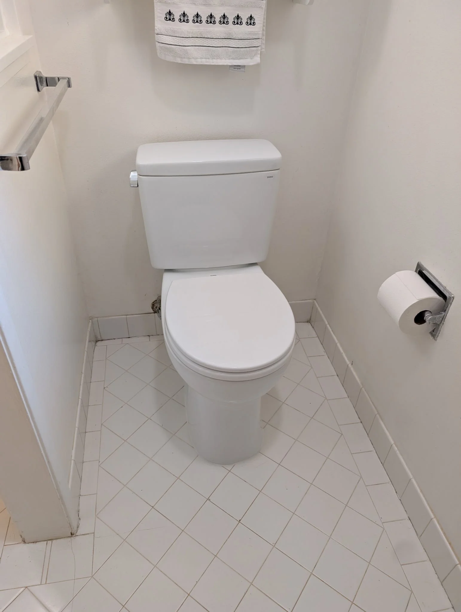 A white toilet in a small bathroom with white tiled floor and a white wall. A towel with a decorative black pattern hangs above the toilet. To the right, there is a toilet paper roll on a wall-mounted holder.