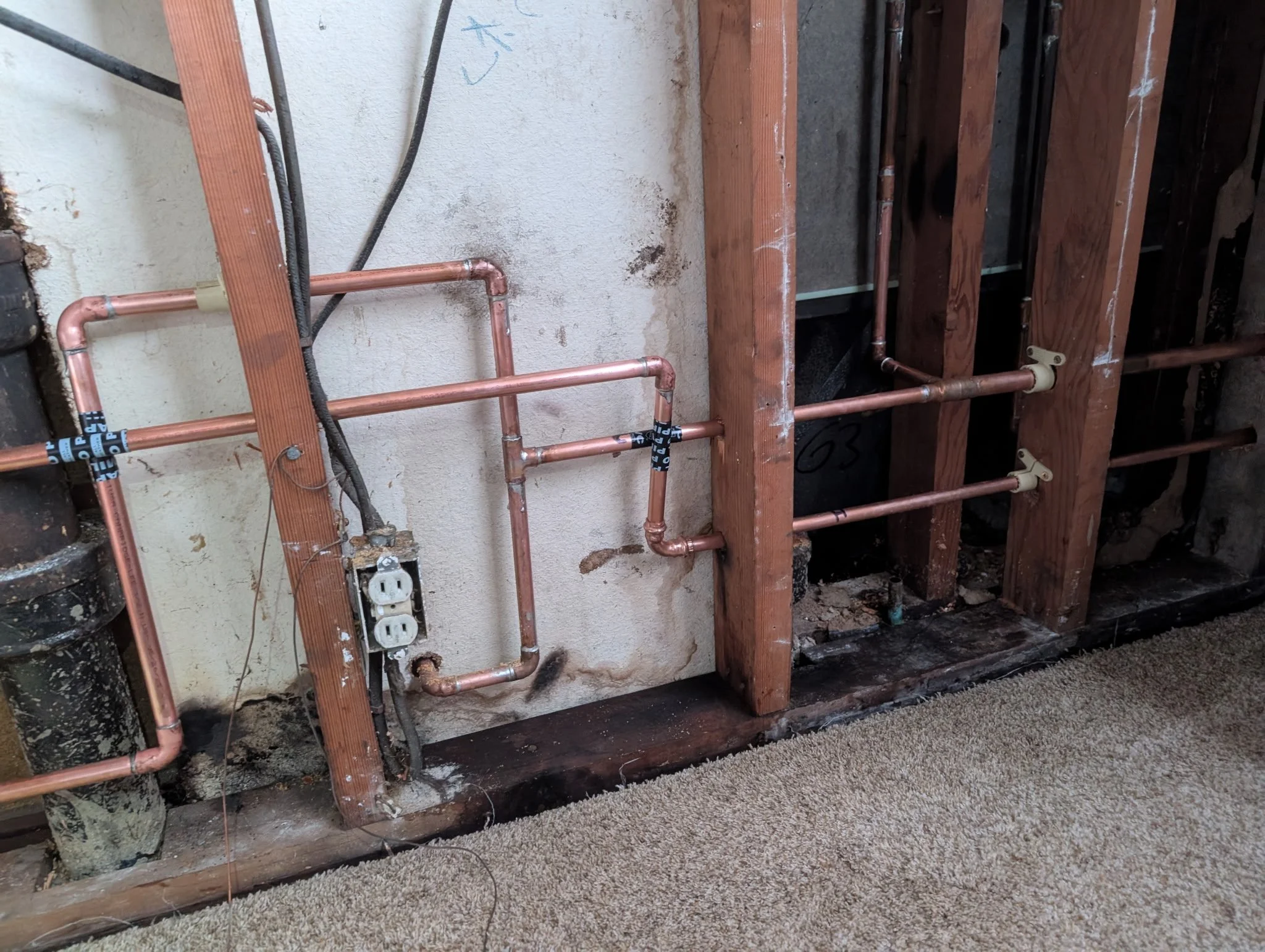 Photo of copper plumbing pipes installed in a wall with wooden framing, with some electrical wiring and an electrical outlet visible.