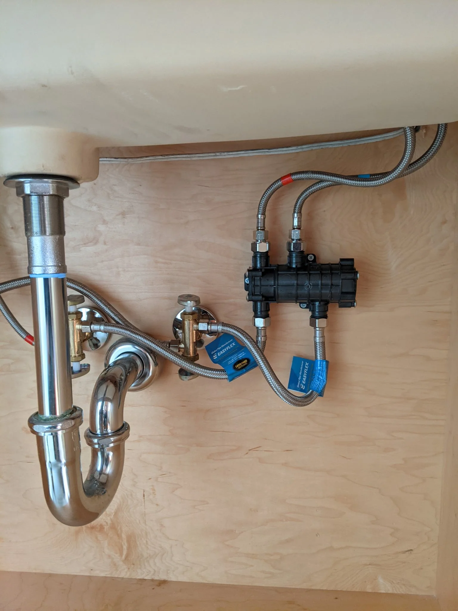 Underneath a sink with exposed plumbing, including a P-trap, flexible hoses connected to a water filtration system, and some blue tags attached to the hoses.