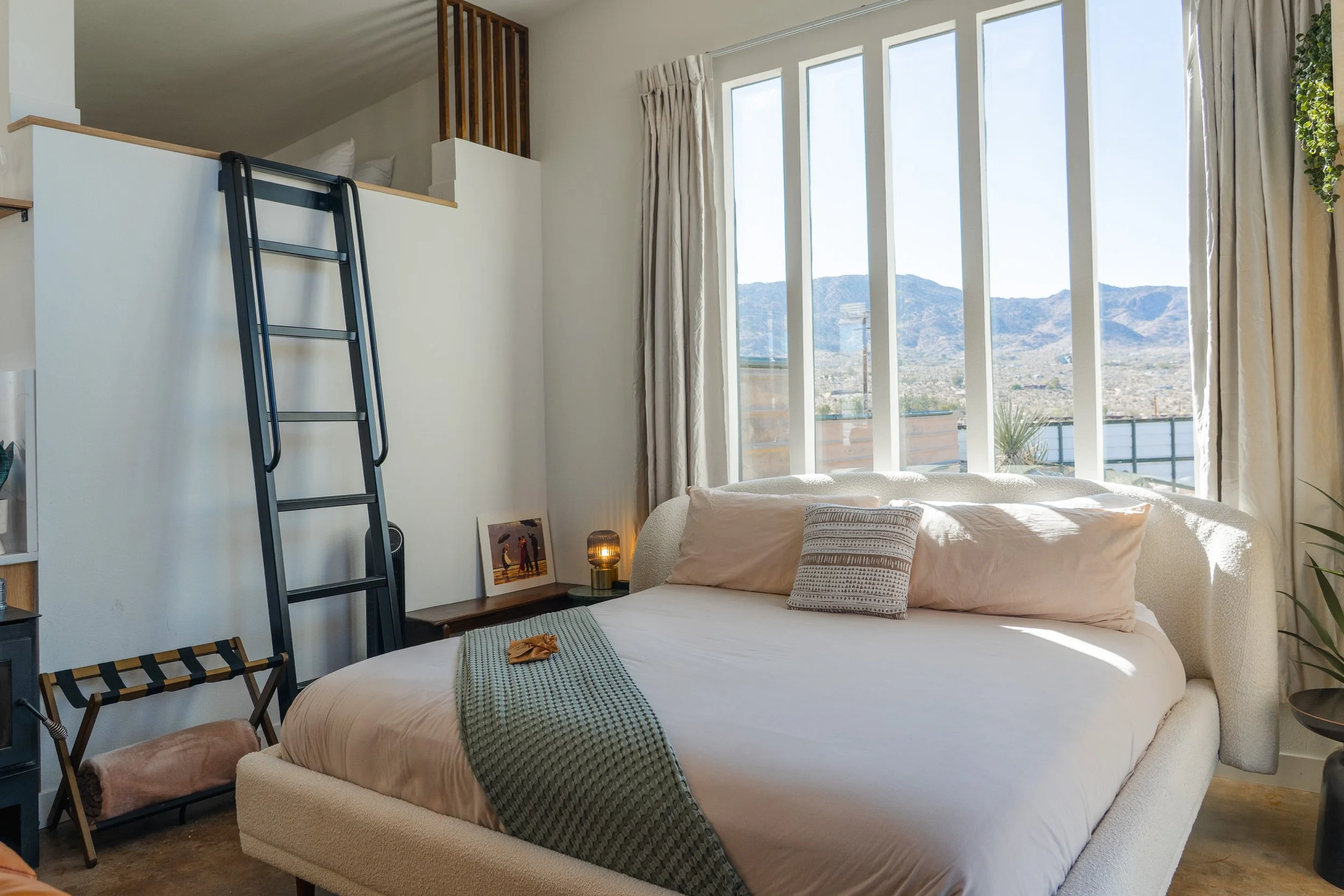 King bed in the open main room at The Atrium, facing tall windows with a desert mountain view, sleeping loft and ladder above