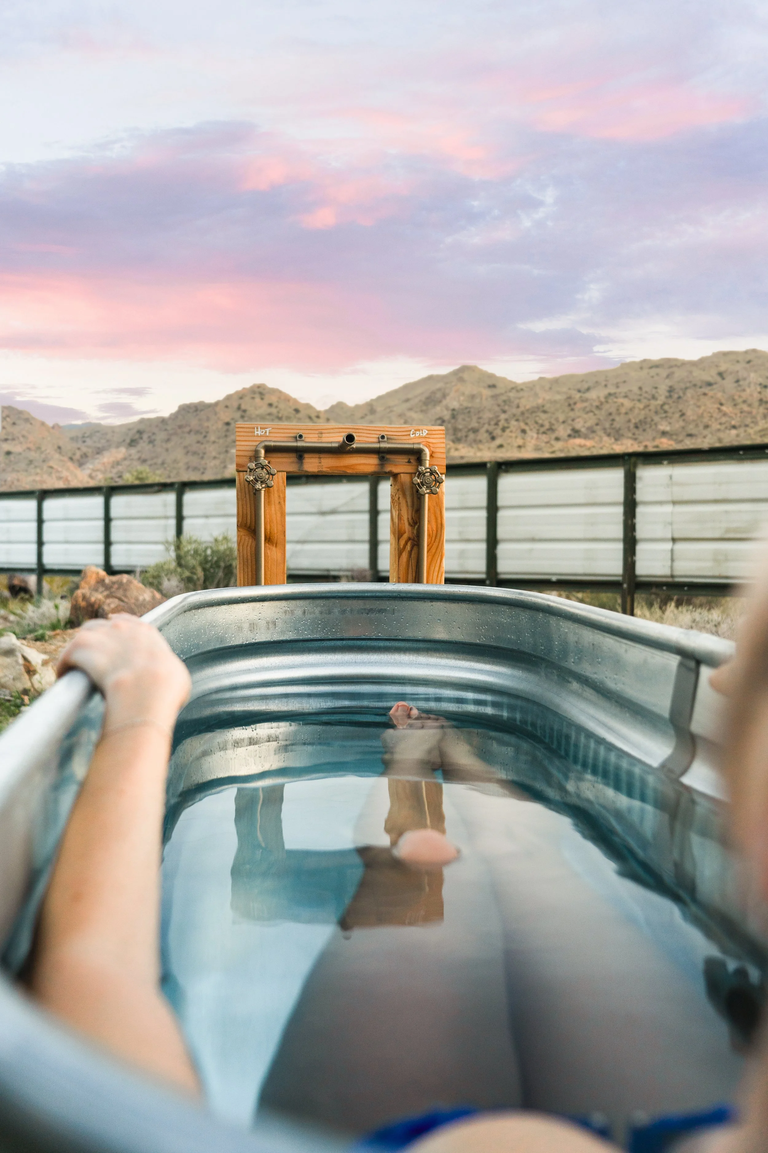 Guest relaxing in a galvanized cowboy soaking tub outdoors at The Atrium, desert mountains and pink sunset sky in the background