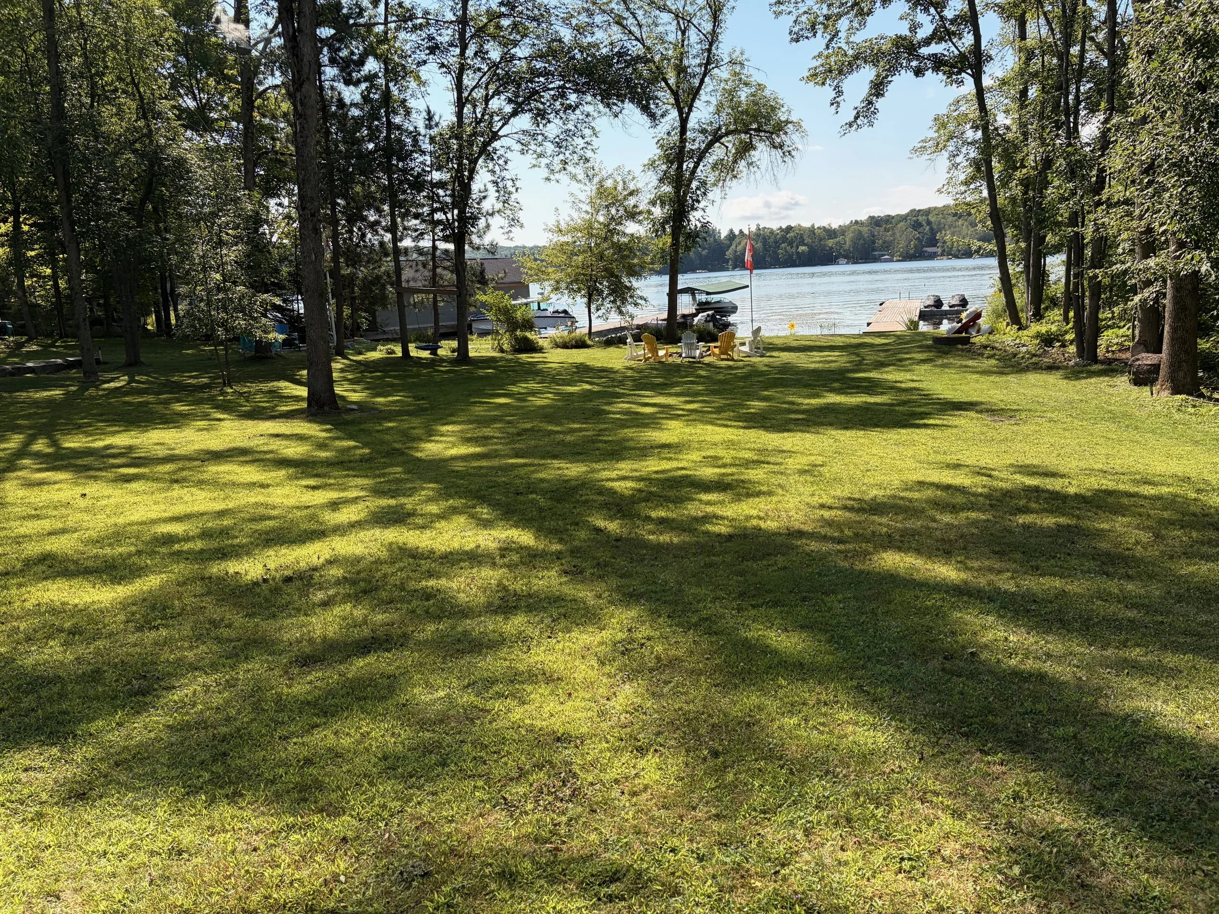 A lakeside scene viewed through trees with green grass in the foreground. There are Adirondack chairs in yellow and white facing the water near a small dock. Several boats are docked, and a Canadian flag is flying. The lake stretches into the distance with wooded land on the opposite shore under a clear, sunny sky.