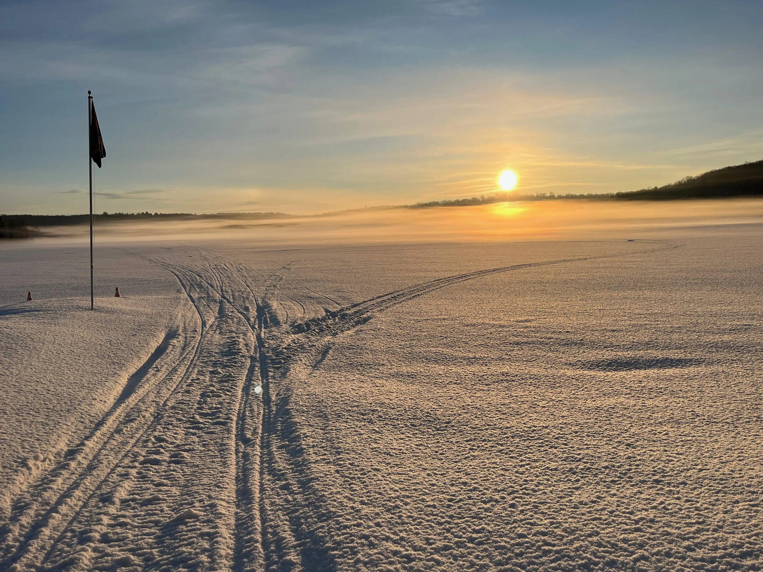 A snow-covered landscape at sunrise with snowmobile tracks and a flag in the foreground, and a hill in the distance.