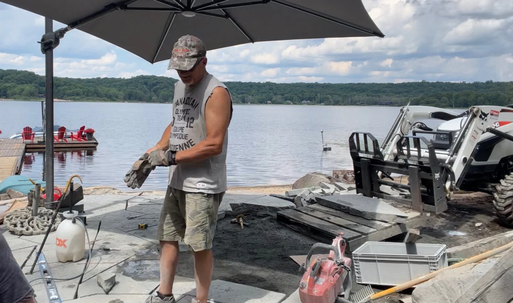 A man working outdoors on a construction project by a lake, with outdoor furniture and a boat visible in the background.