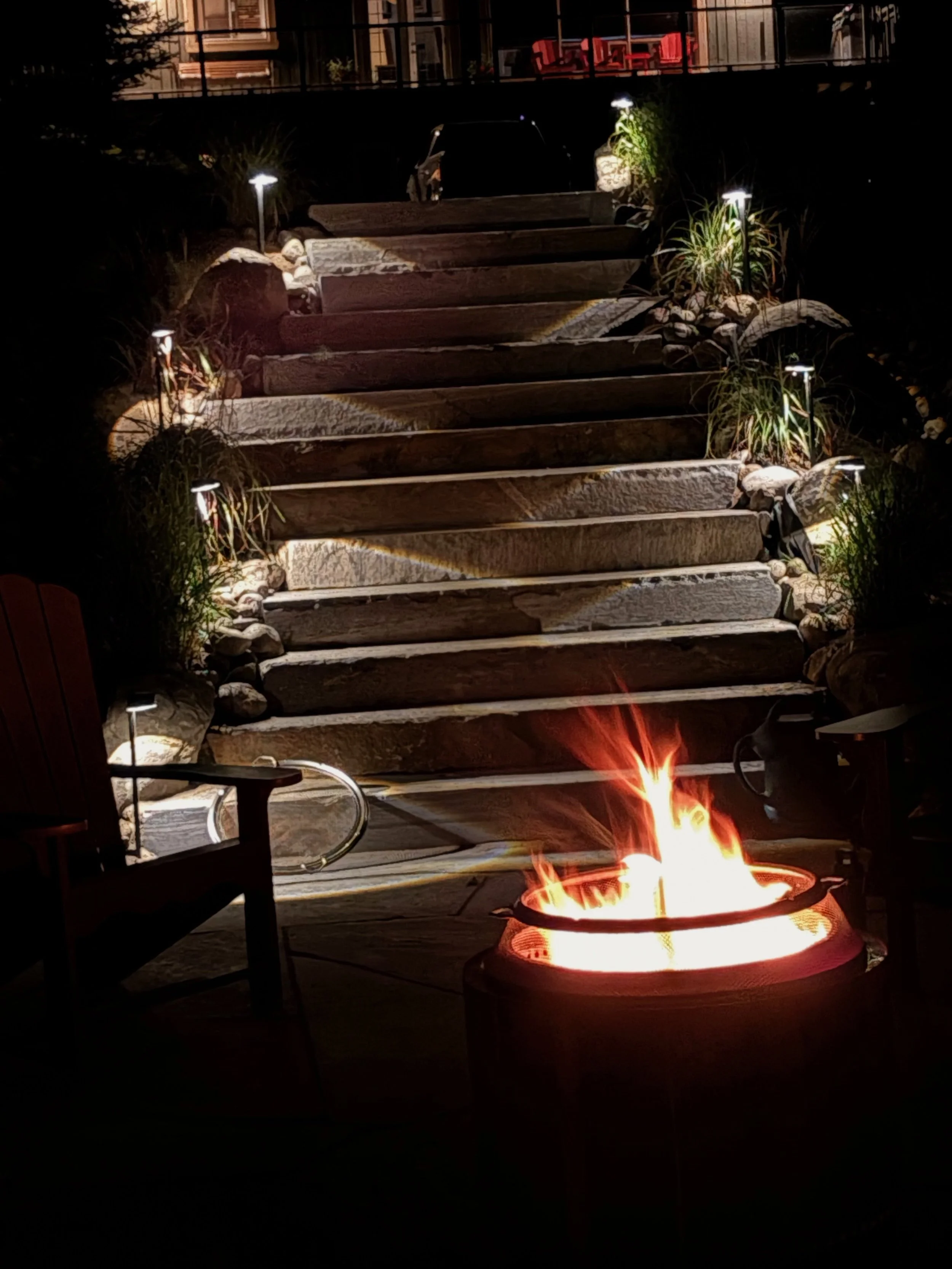 Nighttime scene of a backyard with a fire pit in the foreground, wooden stairs lined with small lights leading up to a patio with outdoor furniture, and a house with lights on in the background.