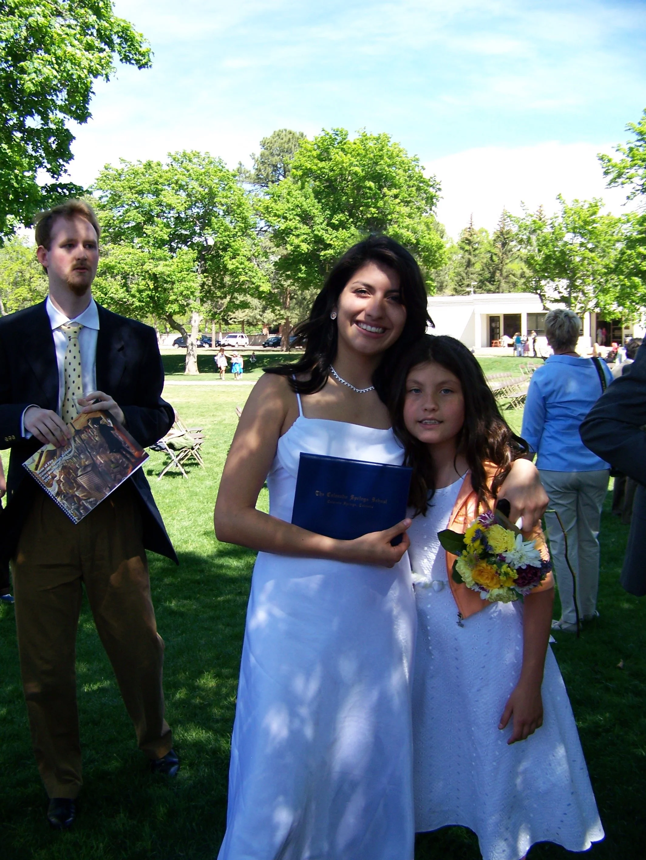 Smile of a woman wearing a white dress holding a bouquet of flowers standing next to a girl in a white dress, with people and trees in the background at an outdoor event.