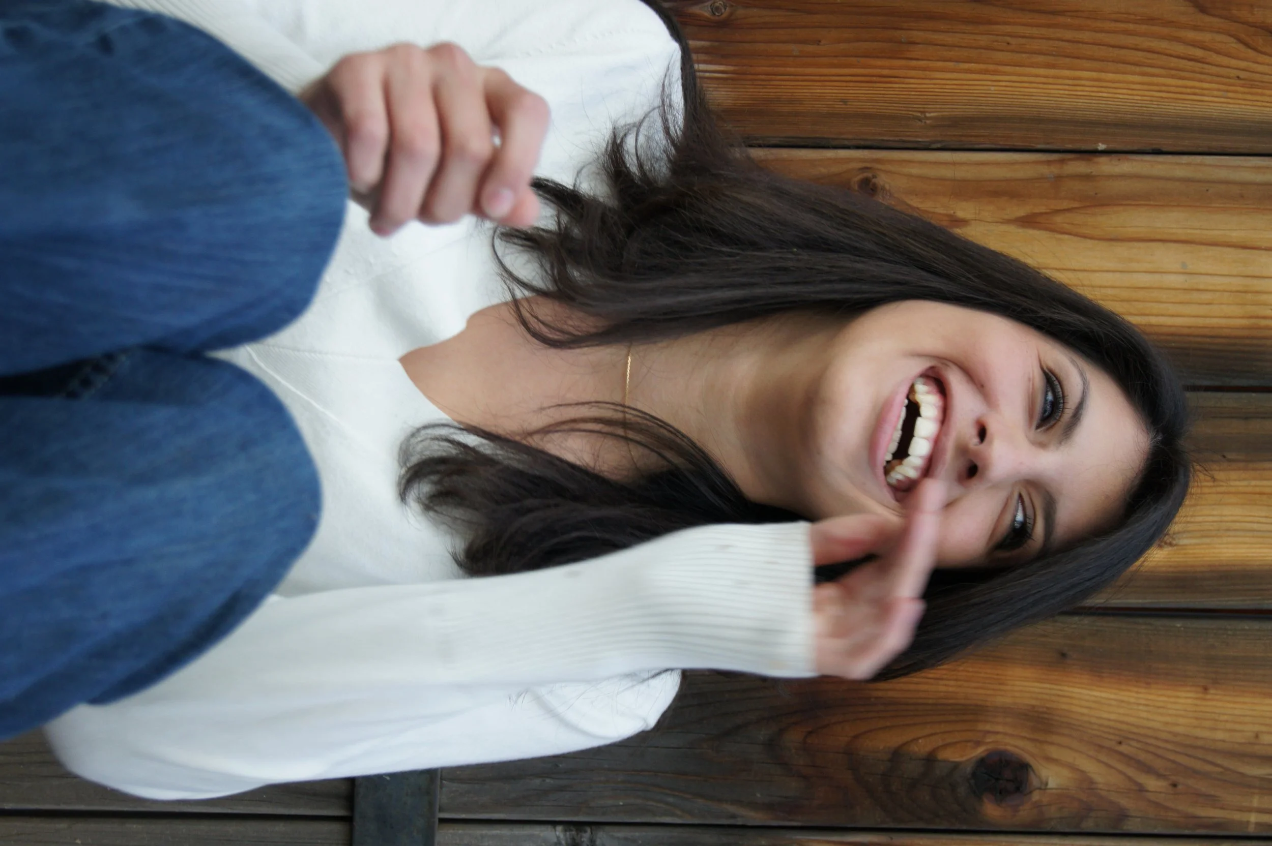 A woman with long dark hair, smiling and laughing, covers her mouth with her hand, wearing a white top, leaning against a wooden wall.