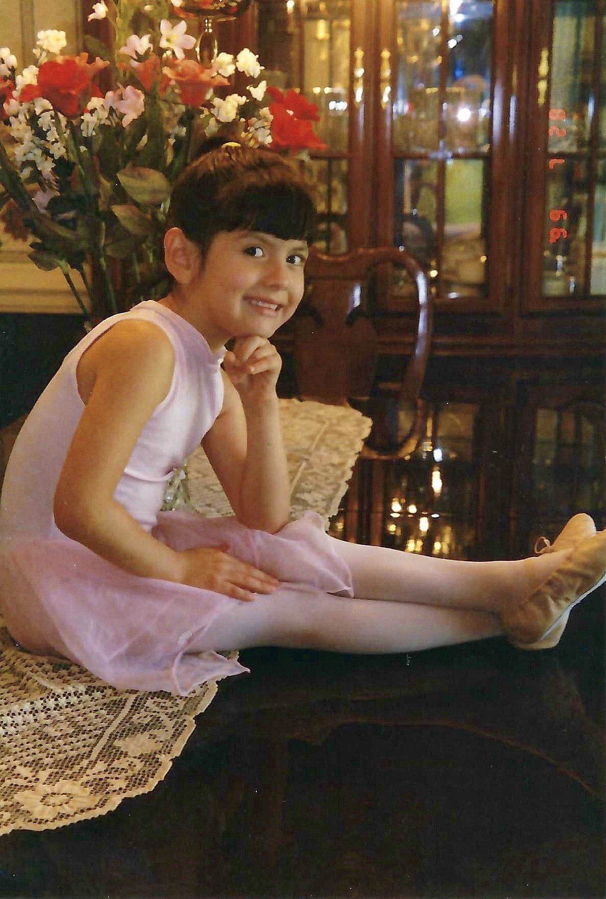 A young girl in a pink dress and tights sitting on a dark wooden table inside a restaurant or cafe. She has short dark hair, smiling, resting her chin on her hand. Behind her, there is a large bouquet of colorful flowers and a wooden cabinet with gla
