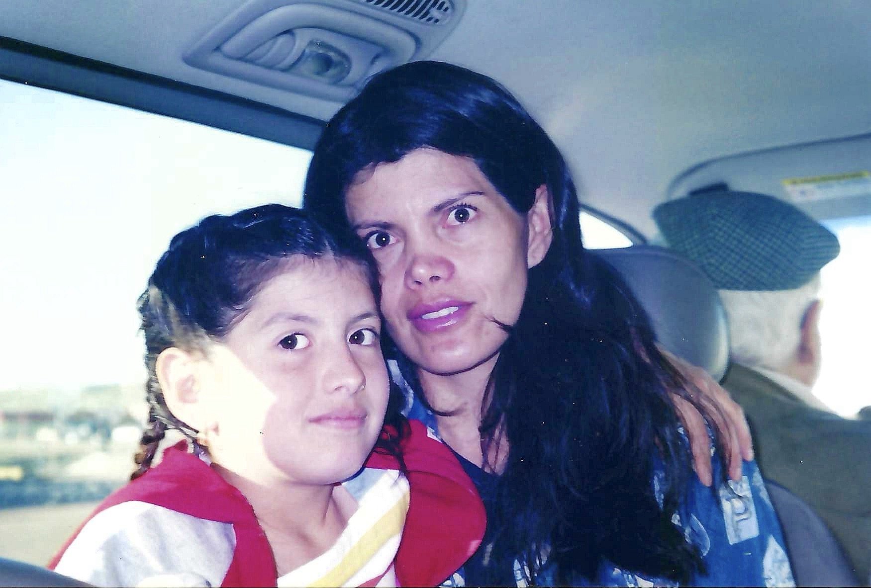 A woman with long dark hair and an intense gaze taking a selfie with a young girl with braided hair and a calm expression, inside a vehicle.