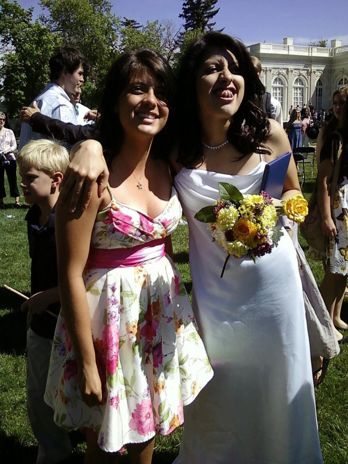 Two women at a wedding, one holding a bouquet of flowers, smiling and posing together outdoors with a crowd and historic building in the background.