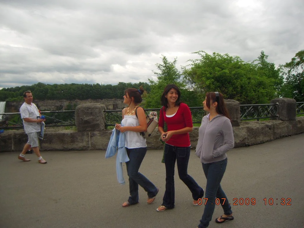 Three women walking and talking on a paved path outdoors, a man standing near a stone railing in the background, with green trees and cloudy sky.