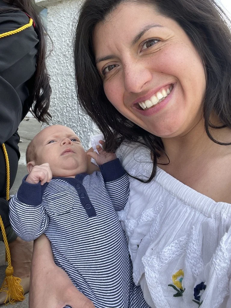A woman smiling while holding a baby outside near a white wall.