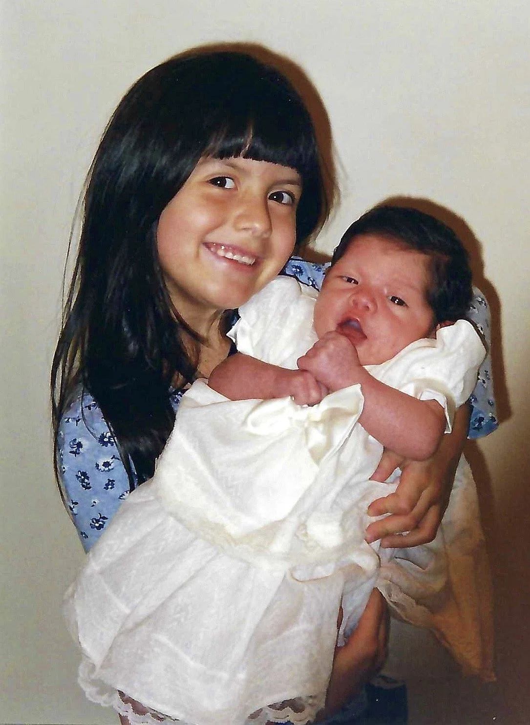 A young girl with long black hair, smiling, holding a newborn baby with dark hair in a white outfit.