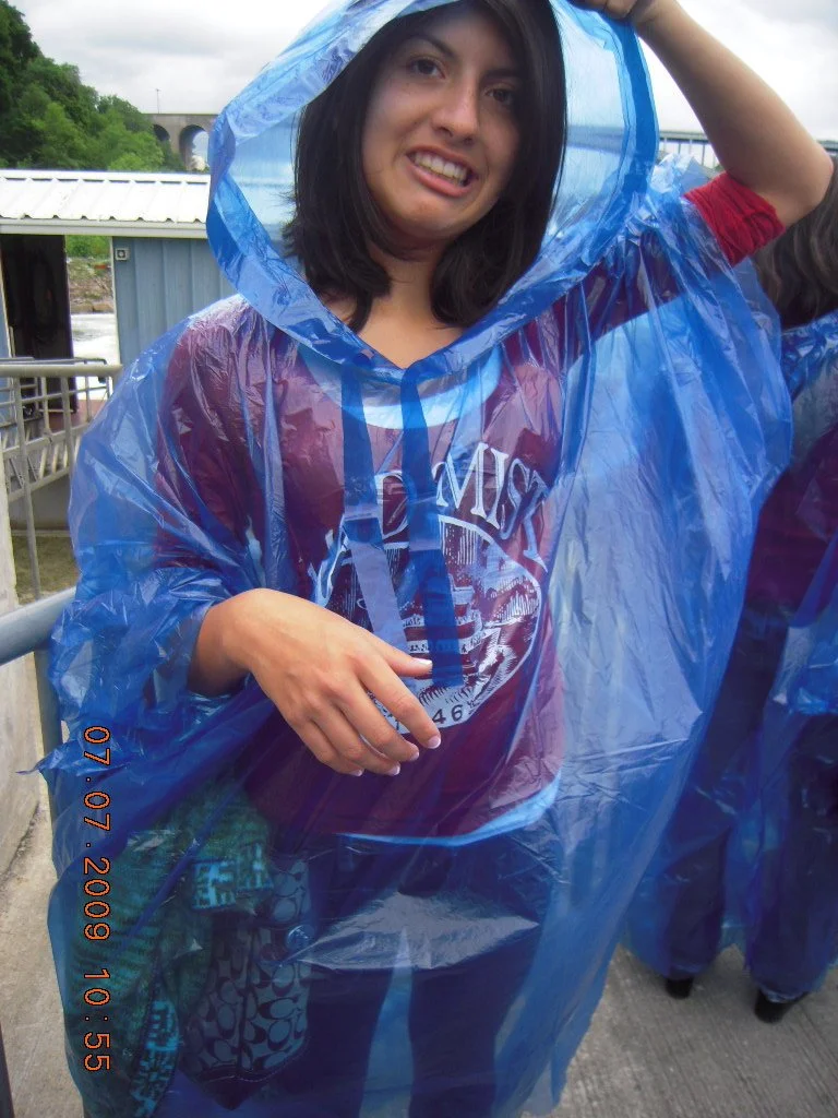 Young woman with dark hair smiling, wearing a maroon T-shirt and a blue rain poncho, standing outdoors near a metal railing and water, possibly during a rainy day.
