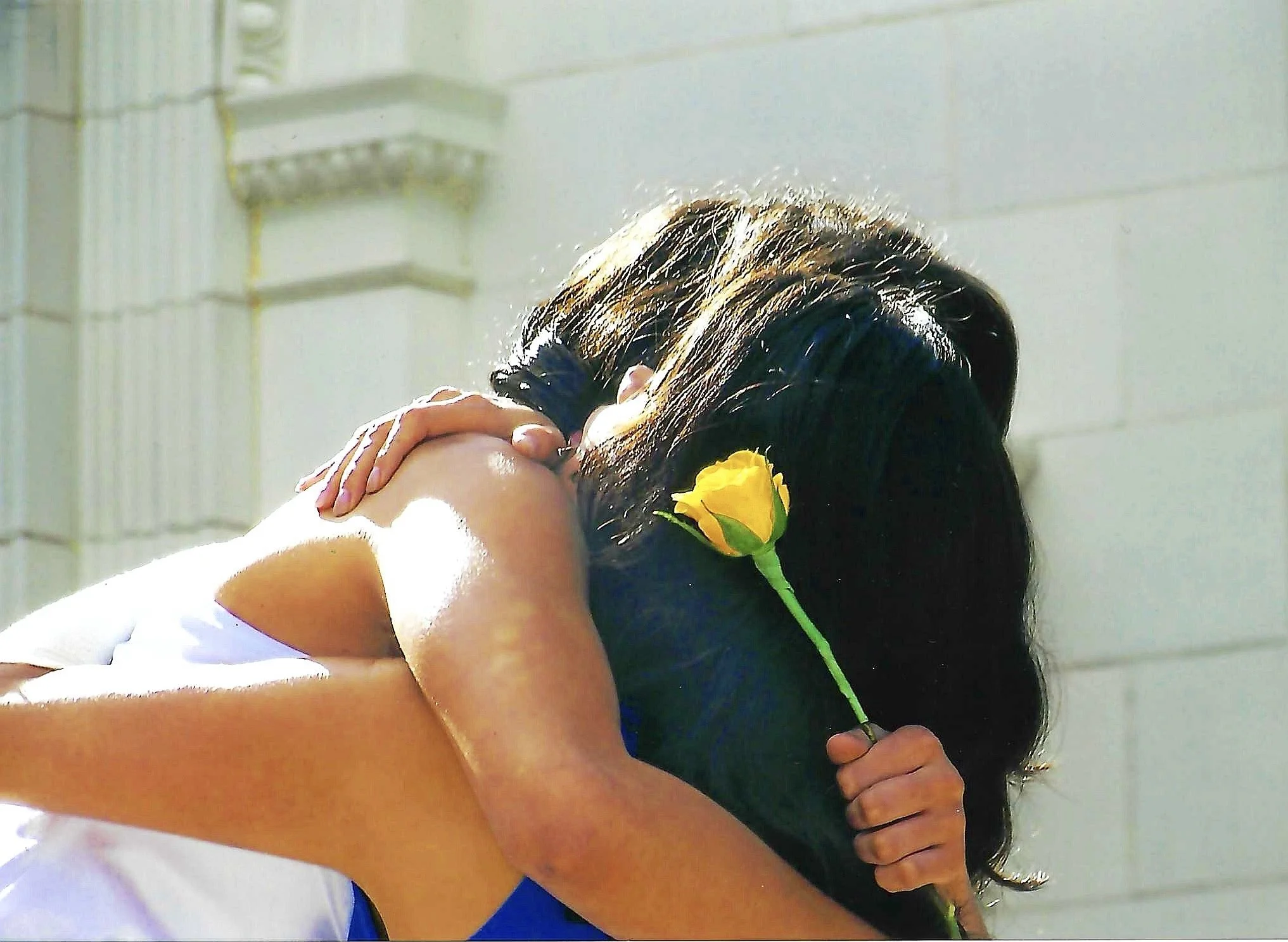 A person embracing another person, holding a yellow flower, with a white tiled wall in the background.