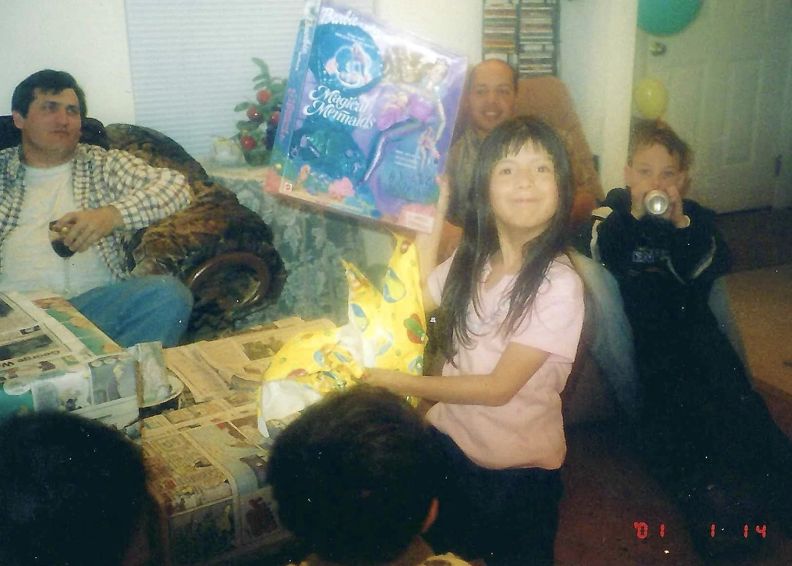A group of people gathered indoors, celebrating with gifts. There is a young girl sitting with a gift bag, and a boy in black holding a soda can. Two adults are seated in the background, one holding a large boxed toy. The room has a small table, a co