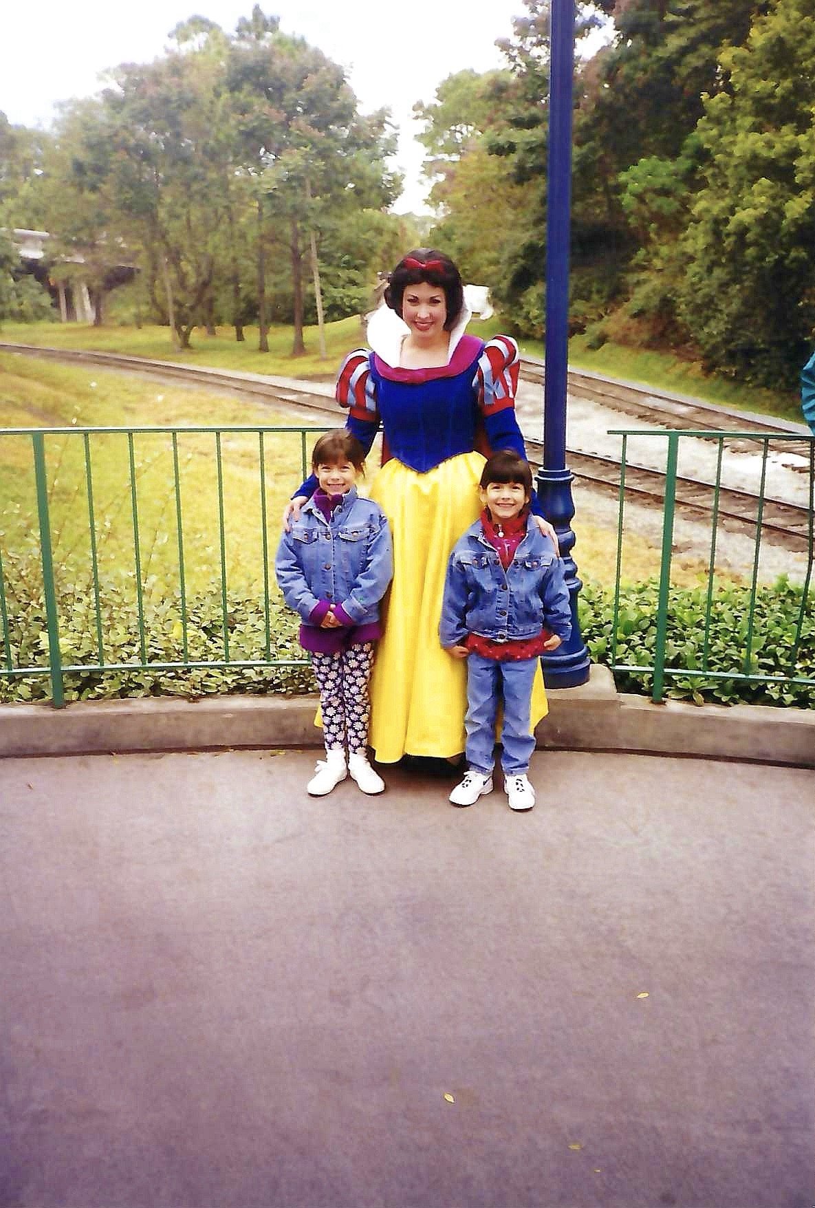 A woman dressed as Snow White standing outdoors with two young girls, all smiling and posing for a photo. The background shows trees, a railway track, and a green fence.