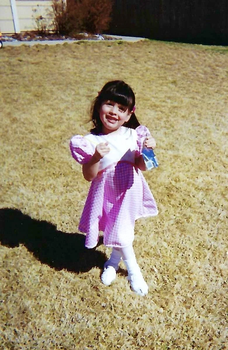 A young girl in a pink checkered dress, white tights, and white shoes, standing on a grassy yard, smiling and pointing at the camera, holding a piece of paper or card.