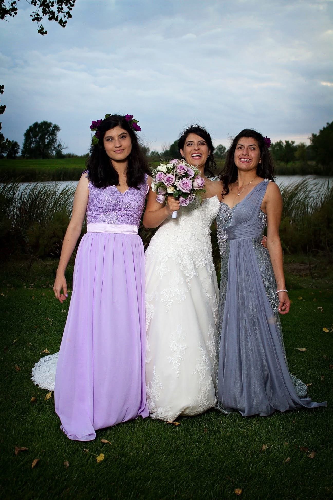 Three women in formal dresses standing outdoors near a lake with trees in the background. The woman in the middle is wearing a white wedding gown and holding a bouquet of purple and white flowers. The other two women are wearing lavender and gray bri