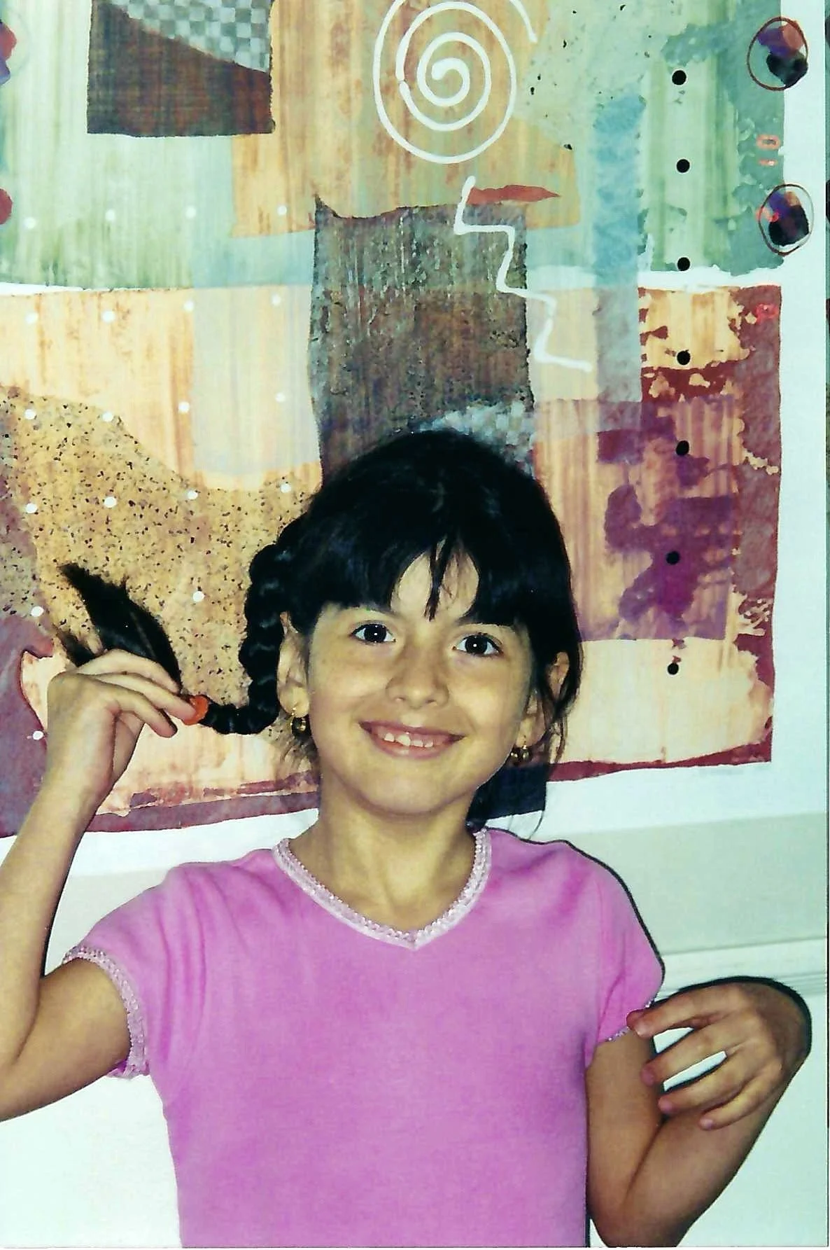 A young girl with dark hair in pigtails, wearing a pink shirt, smiling and holding one of her pigtails. She is standing in front of an abstract, colorful artistic background.