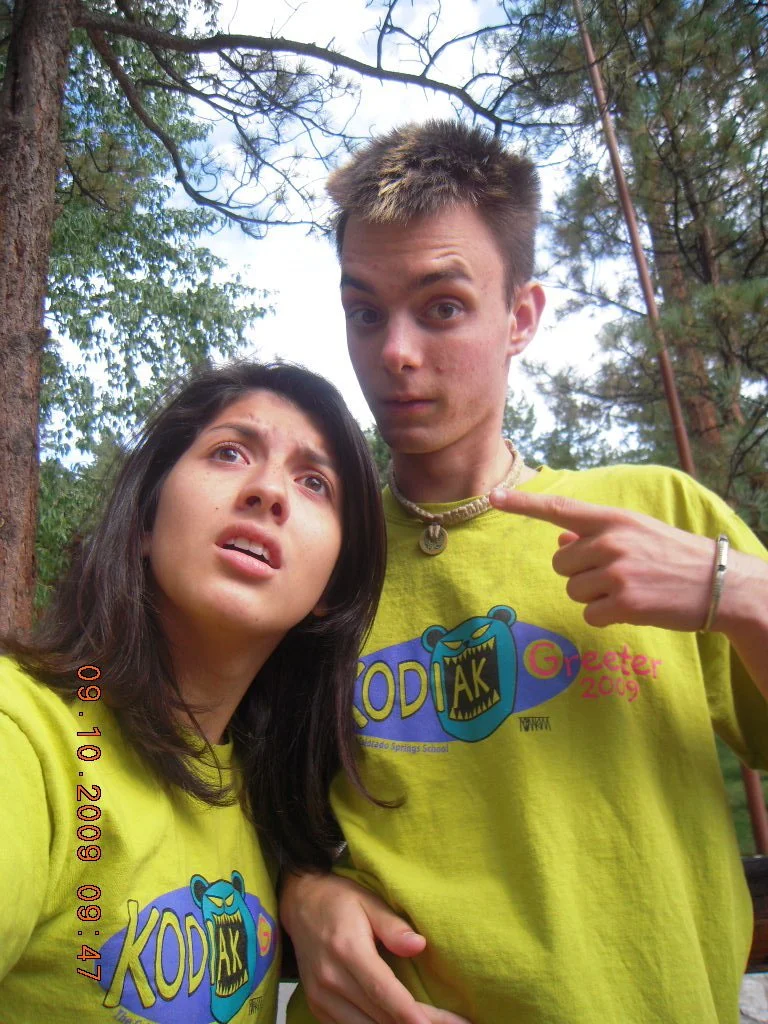 Two teenagers outdoors in a wooded area, making surprised expressions. The girl has dark hair and is wearing a yellow T-shirt with a logo. The boy has short hair, is wearing a similar yellow T-shirt with a logo, and is pointing at himself.