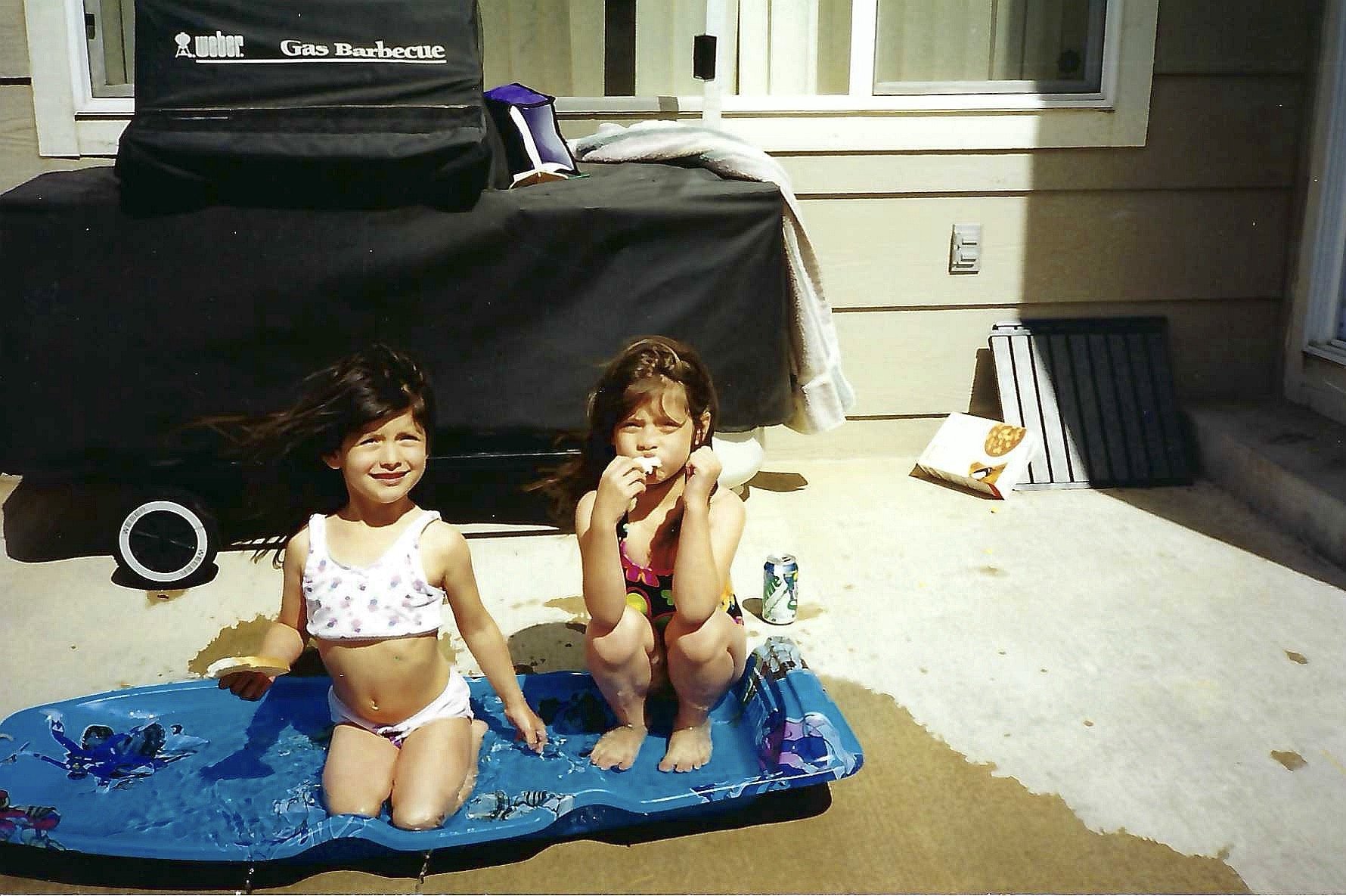 Two young girls sitting in a small inflatable pool on a concrete surface, with a black covered barbecue grill and snacks in the background. One girl is eating, and the other has her hair blowing in the wind.
