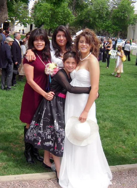 Four women standing together outdoors at a formal event, with trees and other people in the background. One woman is wearing a white dress and holding a white hat, another in a black dress with a pink sash, another in a burgundy dress, and a young gi