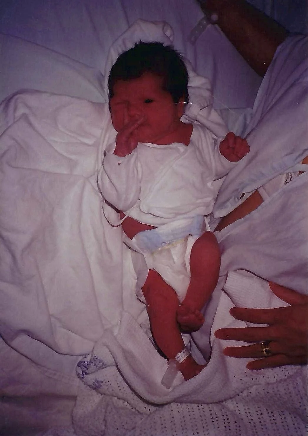 A newborn baby lying on a hospital bed, dressed in a white hospital gown, with an IV attached to one foot, and an adult's hand resting nearby.