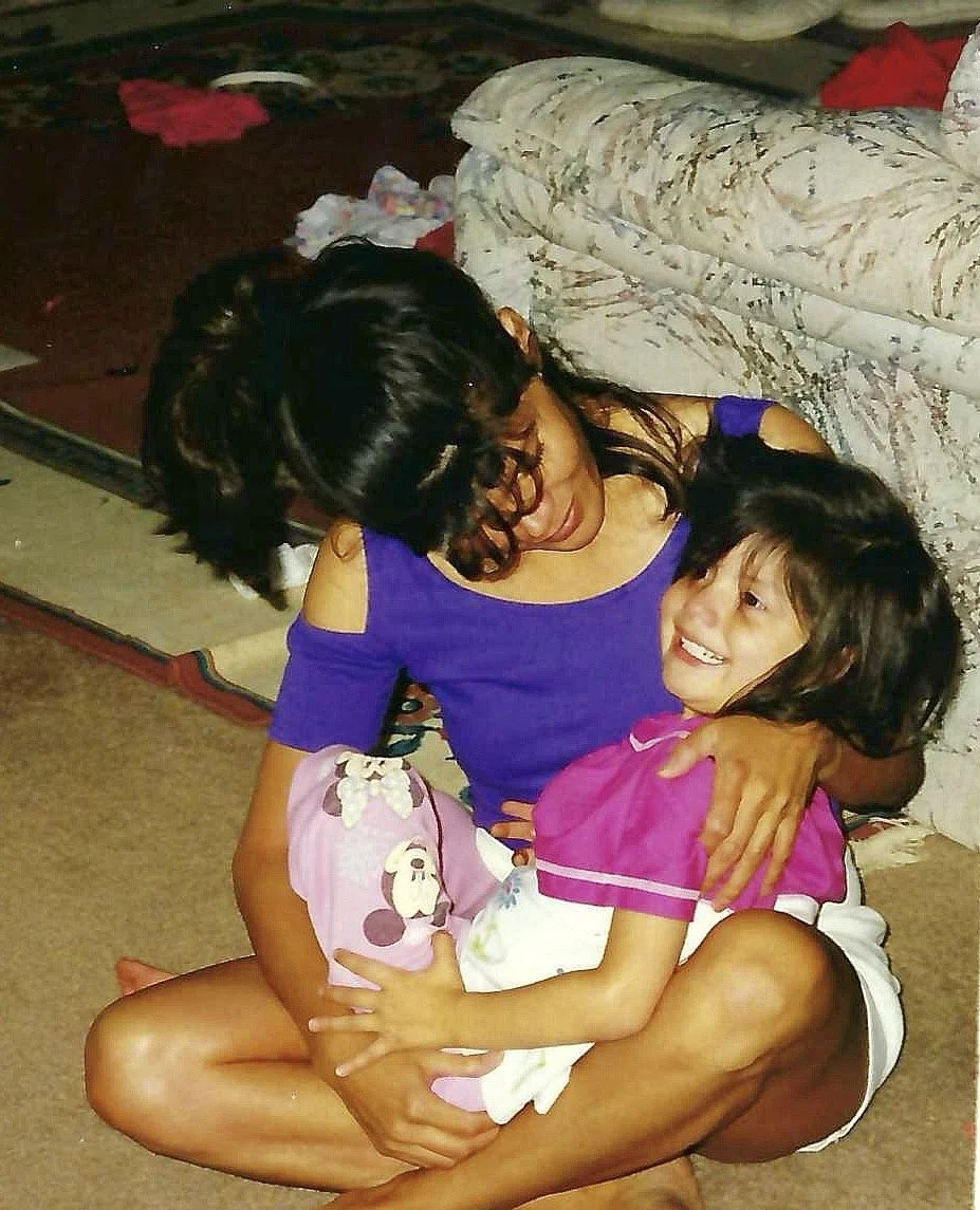 A woman sitting on the floor hugging a young girl, both smiling. The girl is wearing a pink outfit with cartoon character prints. The woman is wearing a purple top with shoulder cutouts. They are indoors near a sofa and a rug.