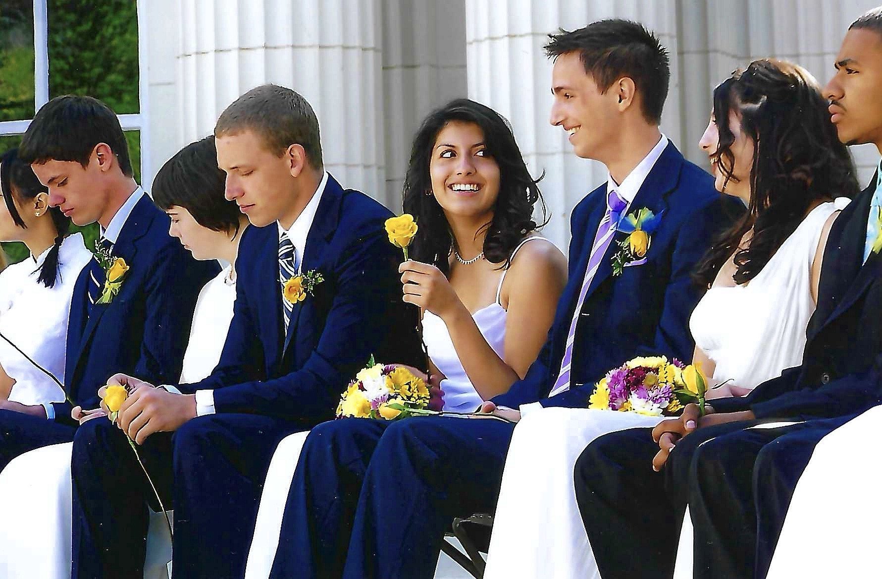 Group of young people dressed in formal attire sitting in a row at an outdoor wedding ceremony, holding yellow and purple flowers.