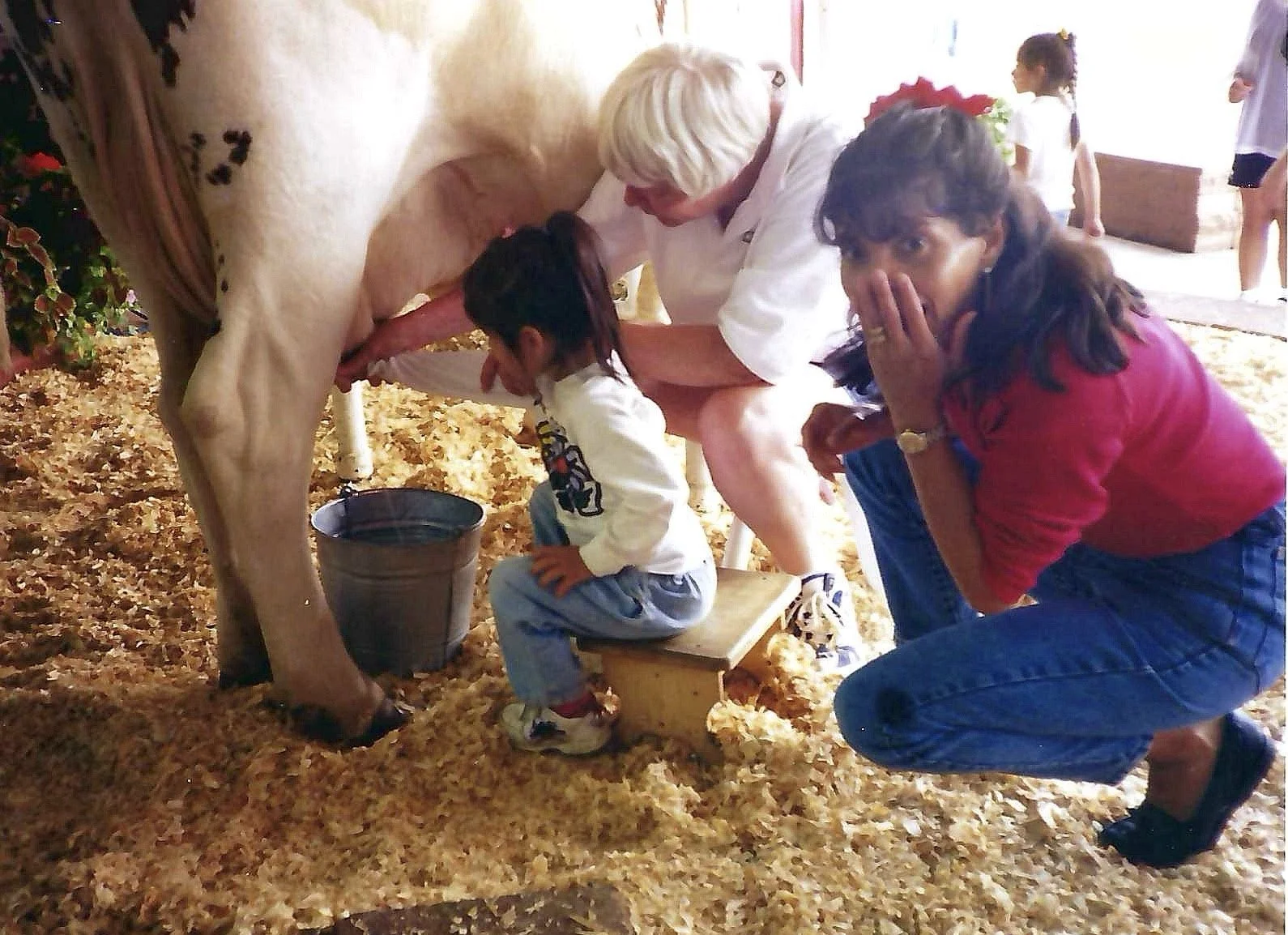 A woman milking a cow while a young girl is seated on a small wooden stool, and a woman in a red shirt covers her mouth with her hand. The scene takes place indoors on a surface covered with straw or bedding materials, with children and adults in the