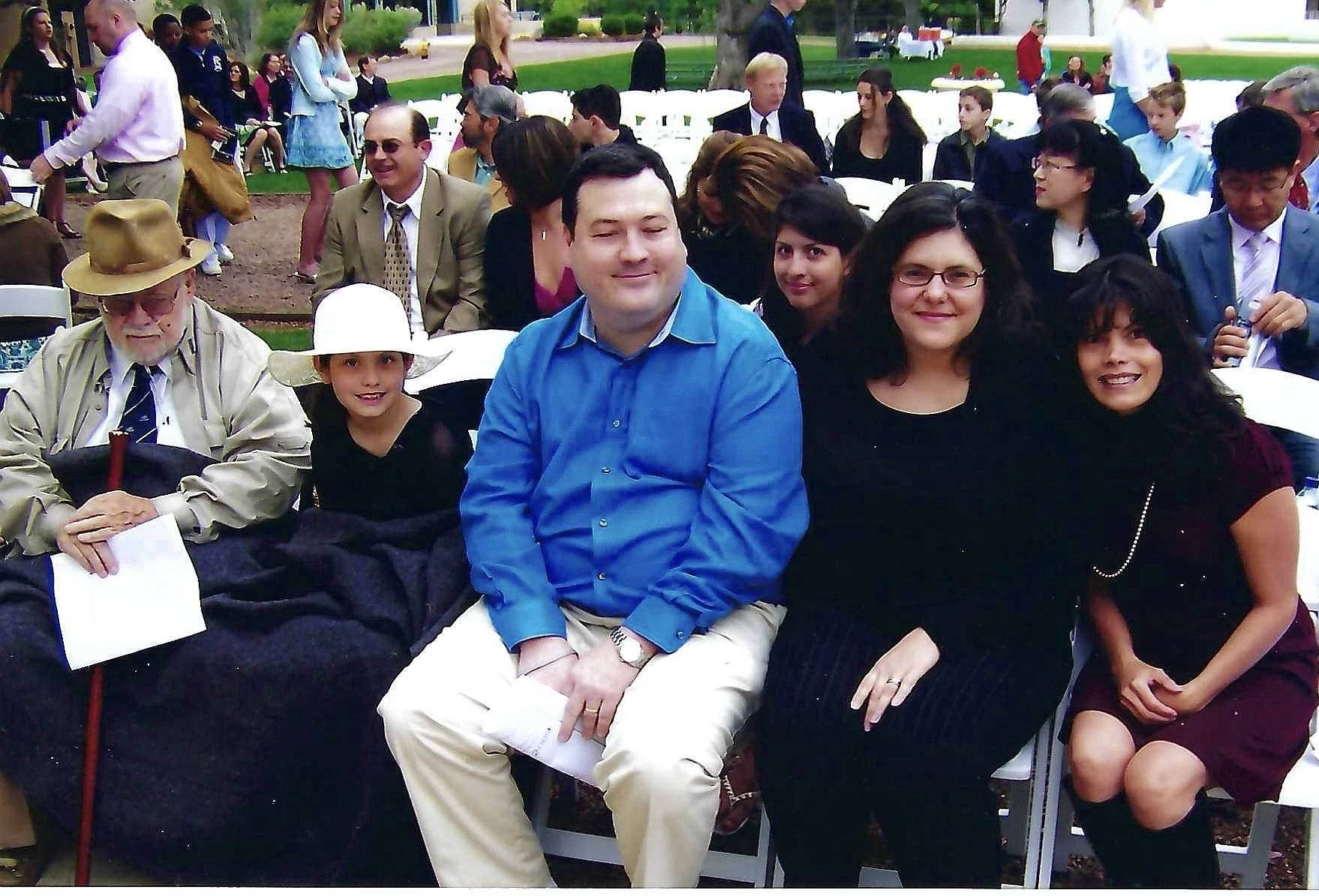 A group of people sitting and standing outdoors at a social gathering or event. Several individuals are smiling and looking at the camera, with some wearing glasses, hats, and formal or casual clothing. There are white chairs and trees in the backgro