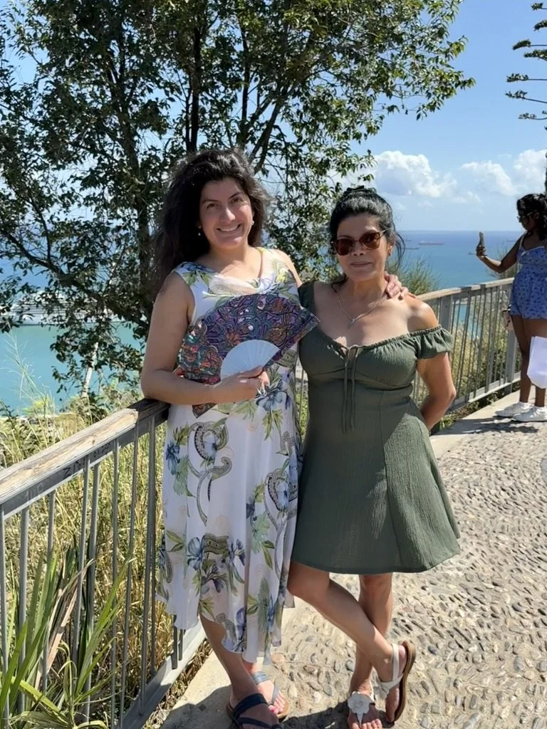 Two women standing outdoors on a sunny day near a railing with a scenic ocean view in the background. One woman is holding a colorful fan, and both are smiling, dressed in summer dresses. A third person is partially visible in the background, taking 