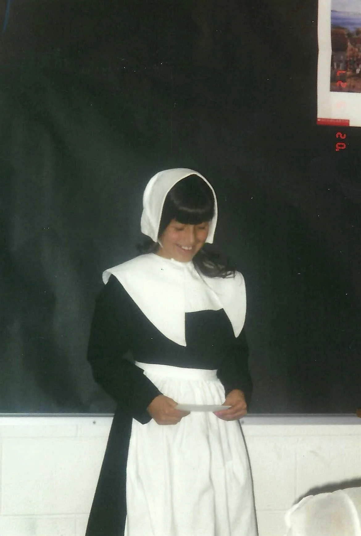 A woman dressed as a nun, wearing a black and white habit, standing in front of a blackboard and smiling while holding a piece of paper.