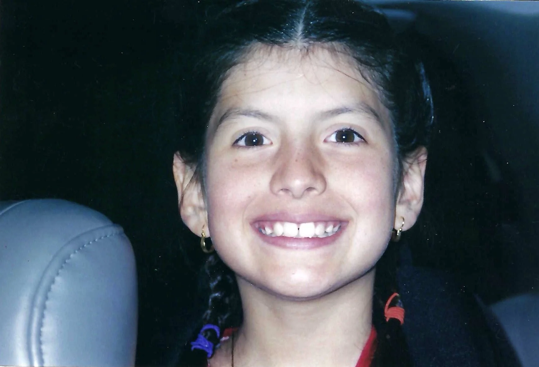 A young girl smiling with braces, wearing earrings, with dark braided hair, and a red shirt, sitting in a car.