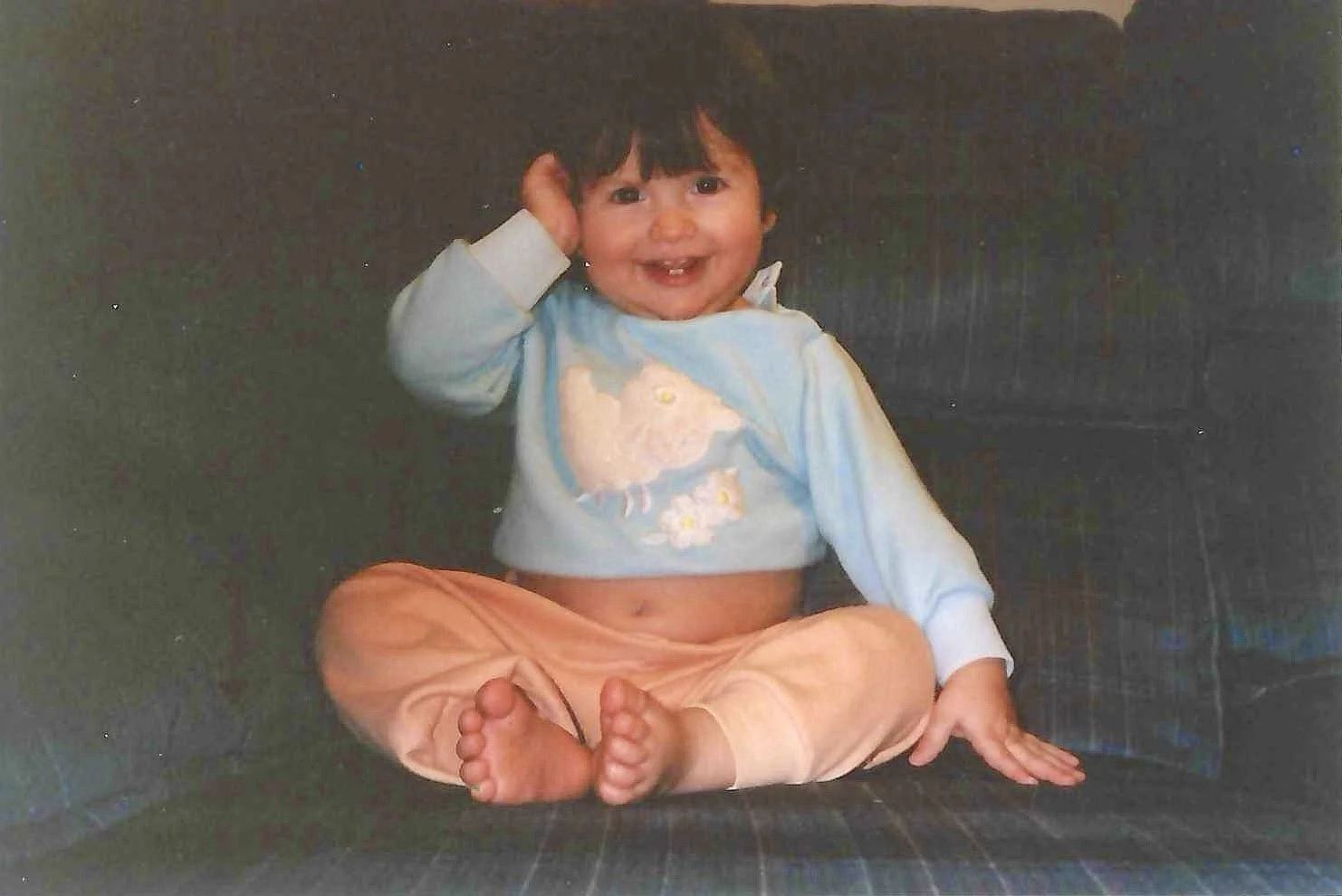 A young child sitting cross-legged on a dark couch, smiling with a missing front tooth, wearing a light blue sweater with a sheep and yellow flowers, and loose orange pants, with one hand touching the side of their head.