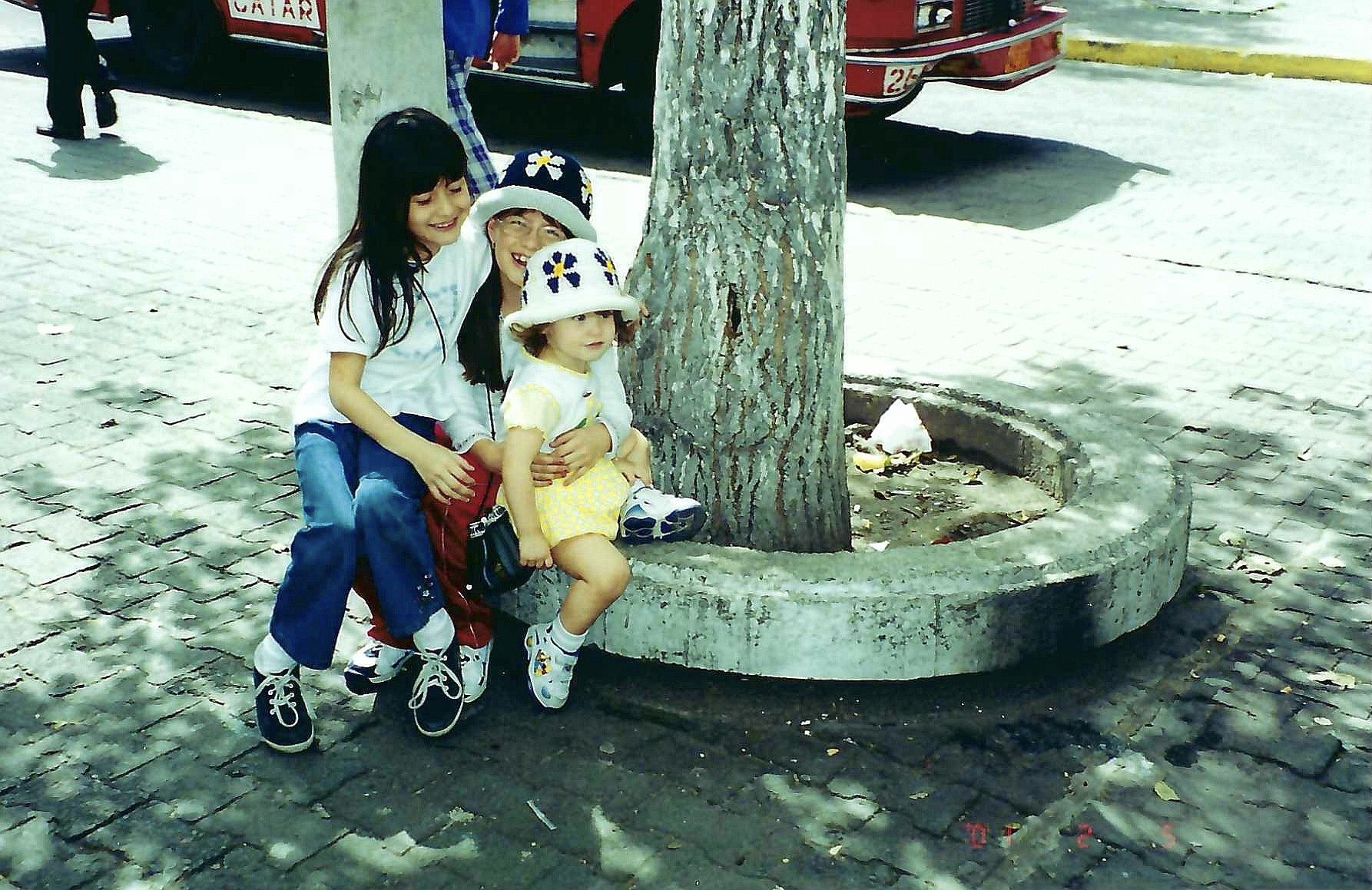 Three children are smiling and sitting on a concrete bench around a tree in a park, with a red bus and a person in the background.