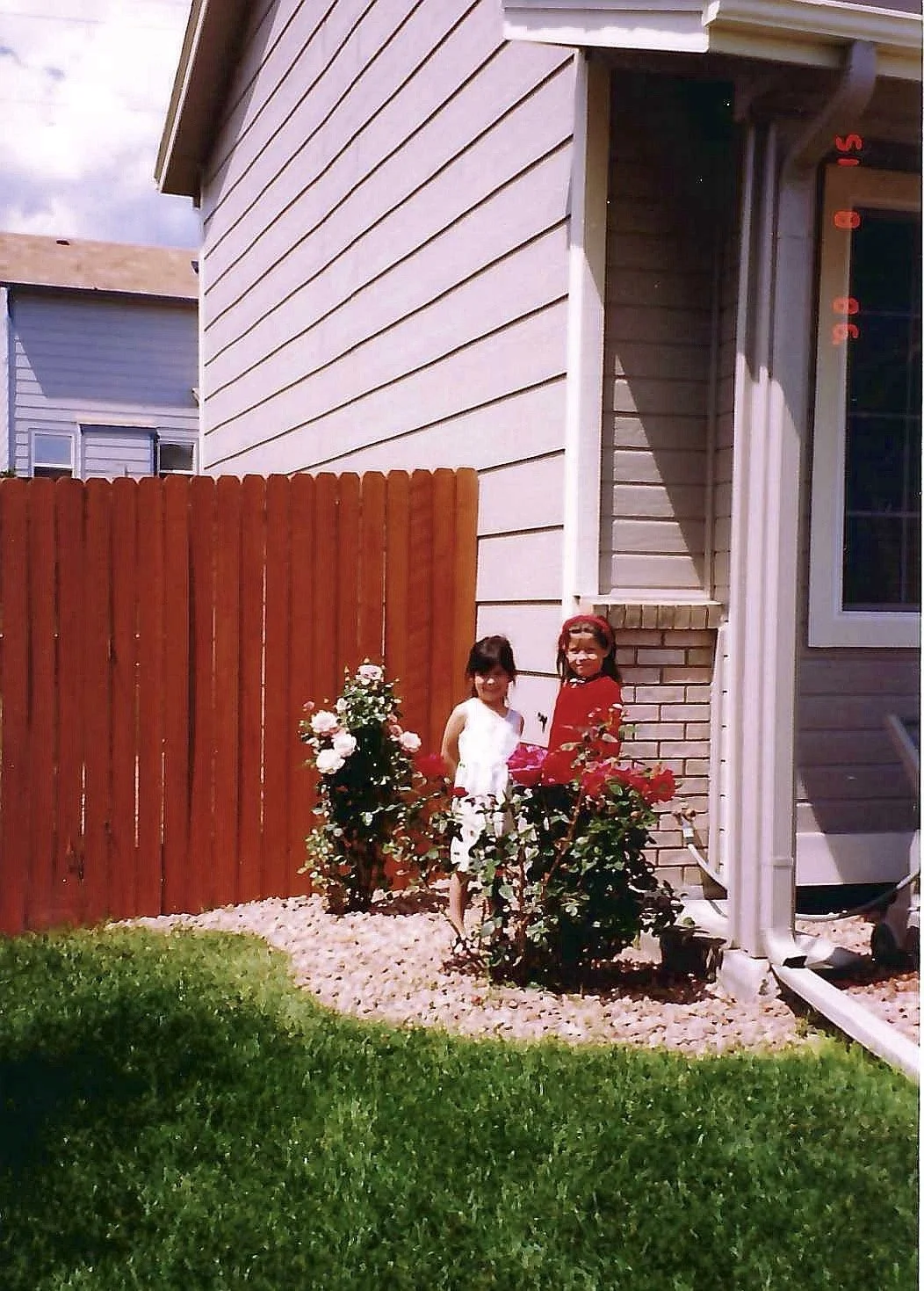 Two young girls standing in a small flower garden with pink and white roses, next to a house with beige siding and a red wooden fence.