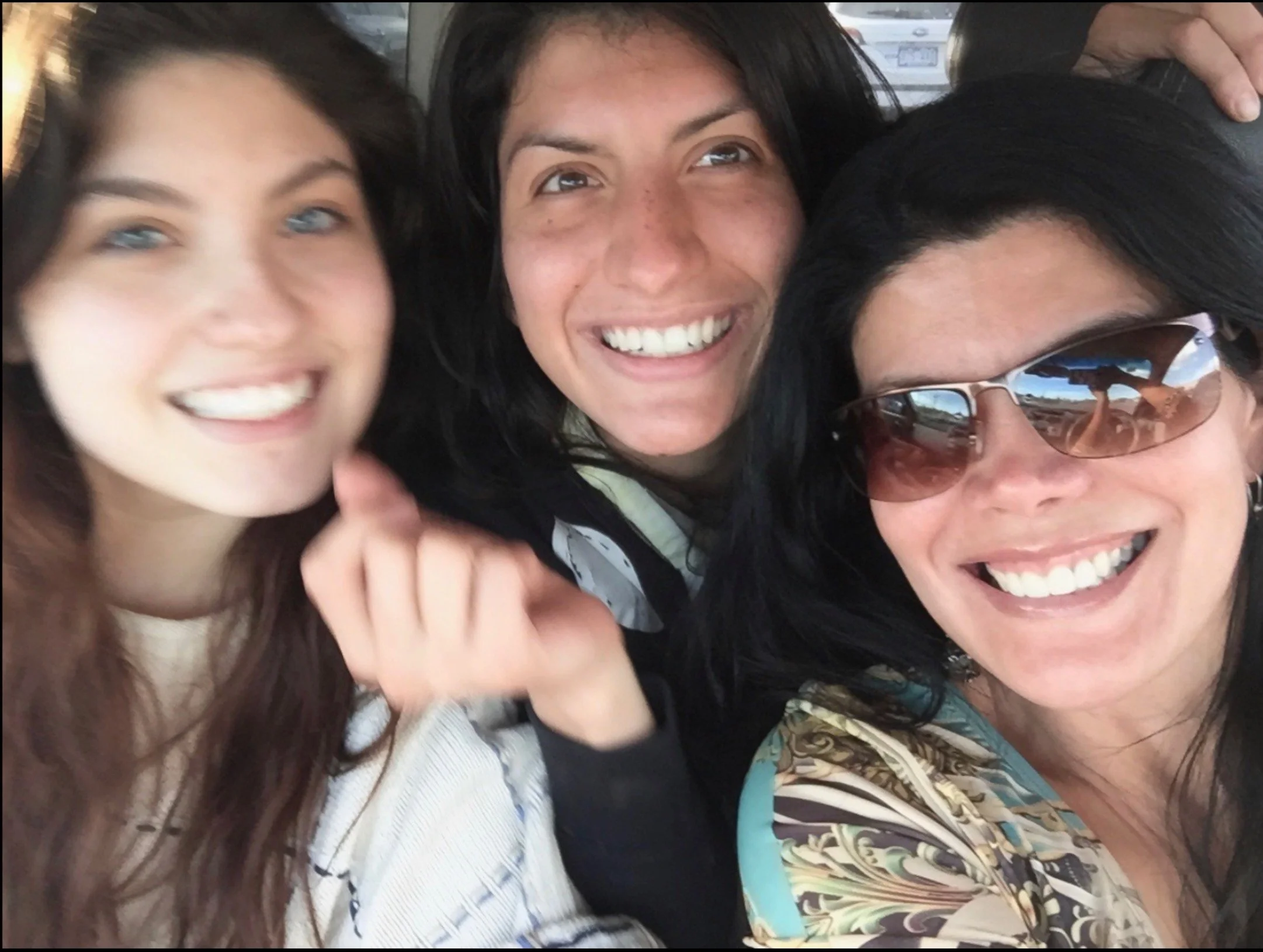 Three women smiling closely together, two with dark hair and one with brown hair, in a car with one wearing sunglasses.