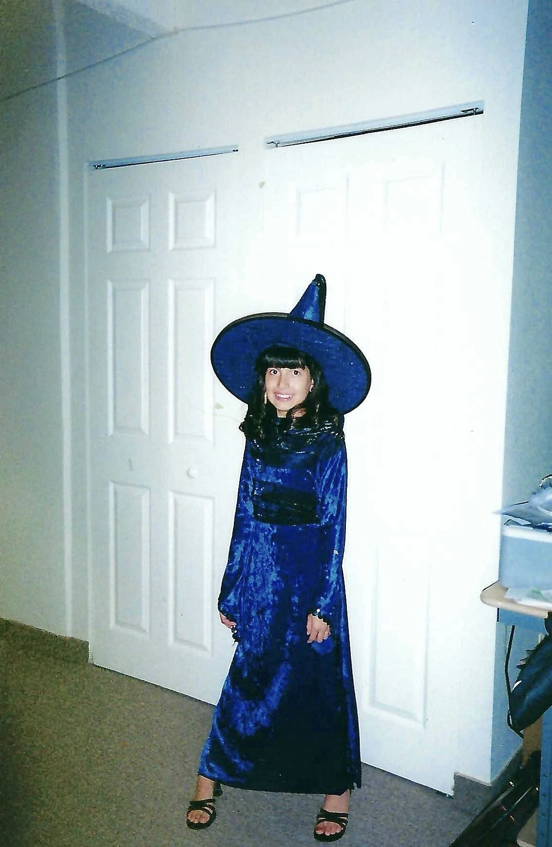 A young girl dressed as a witch in a blue velvet dress and a matching tall hat, standing in front of white double doors, smiling at the camera.
