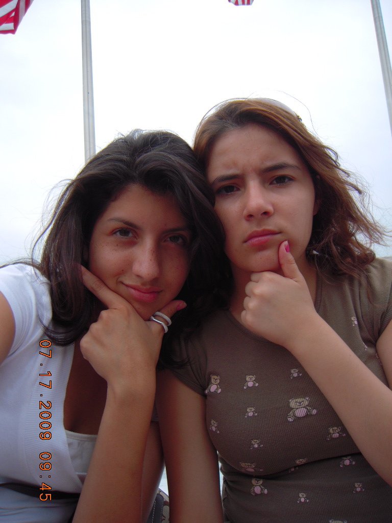 Two young women posing for a selfie outdoors during the daytime. Both have thoughtful expressions, with one resting her chin on her hand. The woman on the left has dark, wavy hair and is wearing a white top. The woman on the right has light brown, cu