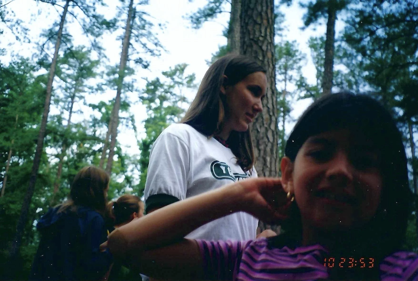 Group of people, including women and children, gathered outdoors in a wooded area with tall trees, engaging in an activity or social event.