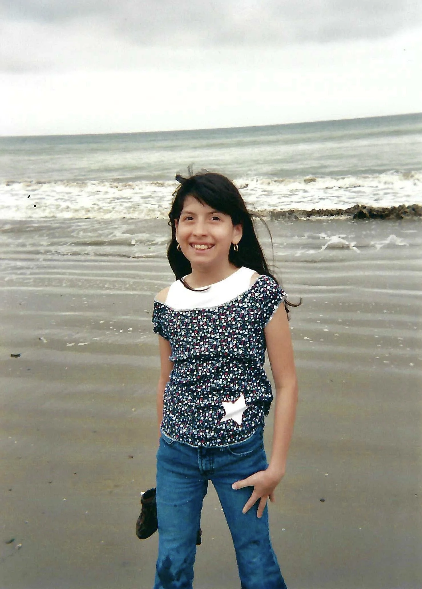 A young girl with dark hair, wearing a floral shirt and jeans, smiling at the camera on a beach with ocean waves in the background.