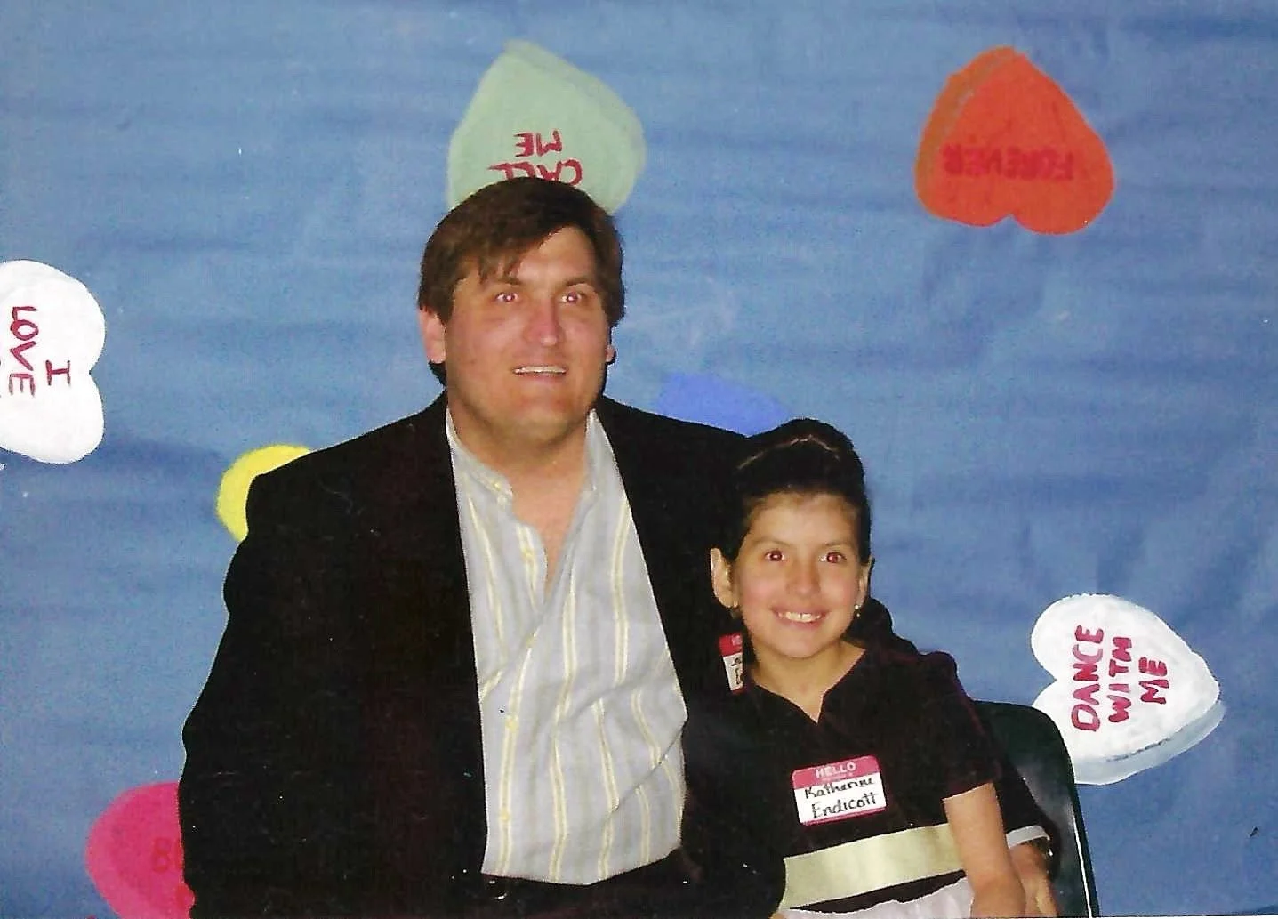 A man and a young girl smiling and sitting together in front of a blue backdrop decorated with colorful paper hearts with messages.