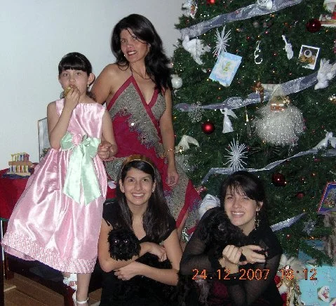 Four women and a young girl in front of a decorated Christmas tree, with two women holding puppies, celebrating Christmas.