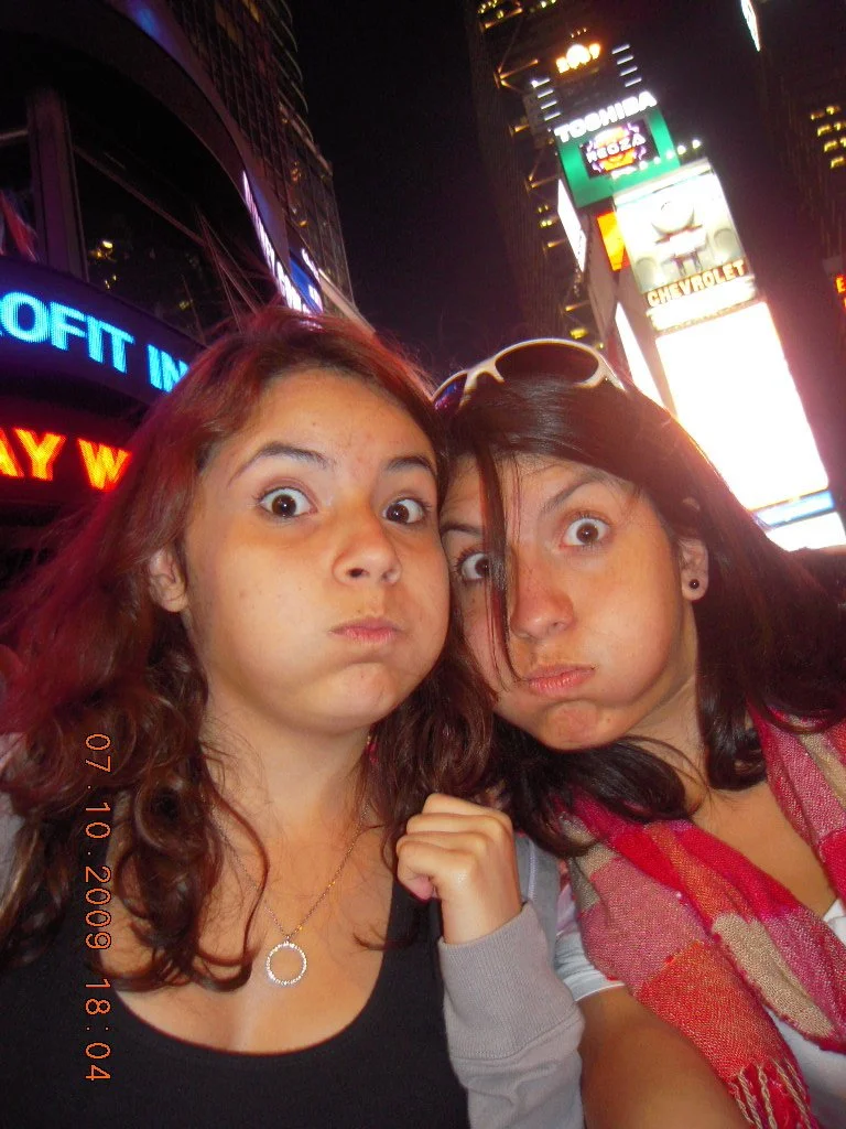 Two young women with surprised facial expressions taking a selfie at night in Times Square, New York City.