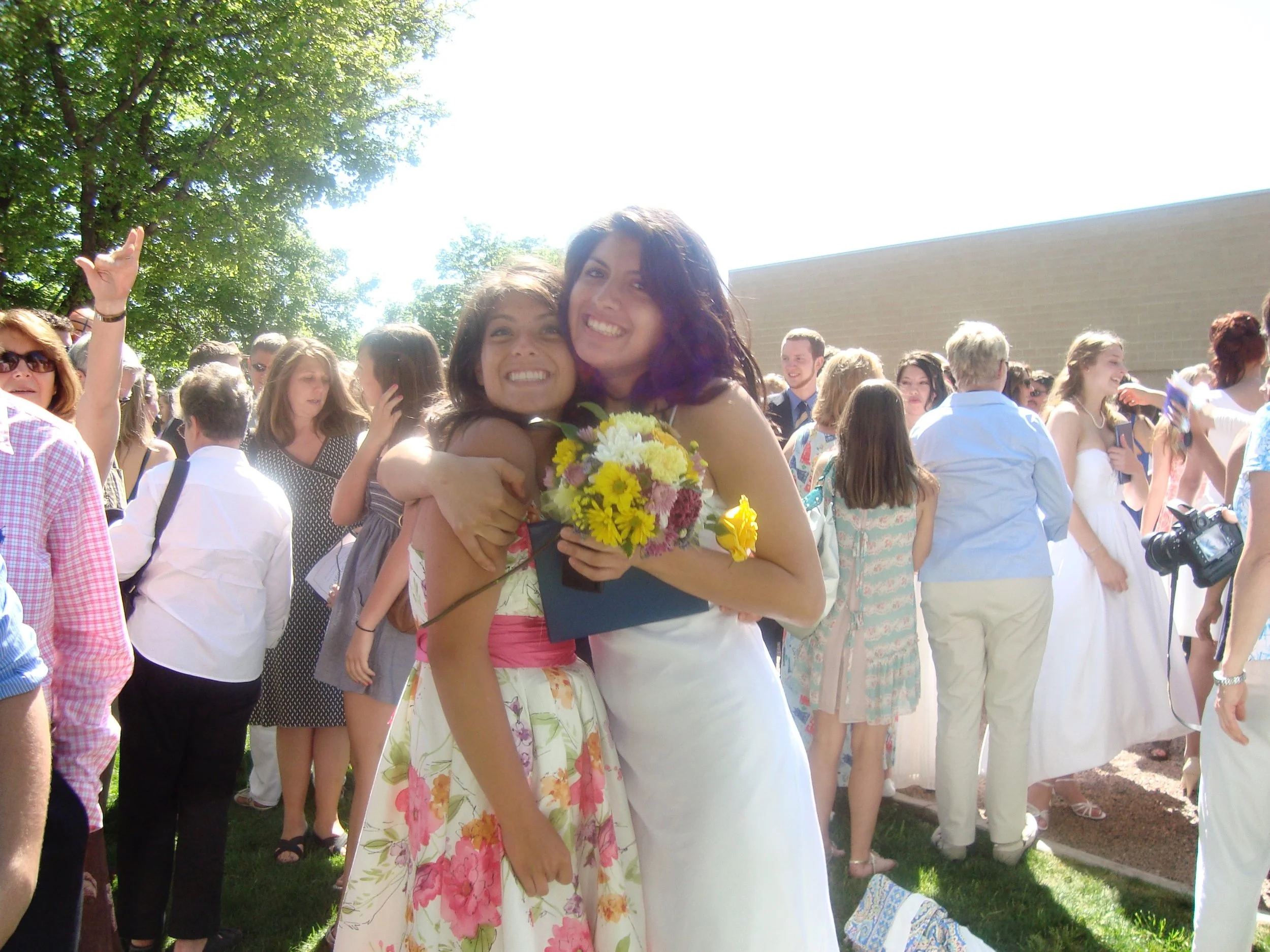 Two women hugging at an outdoor event with many people, trees, and a building in the background on a sunny day.